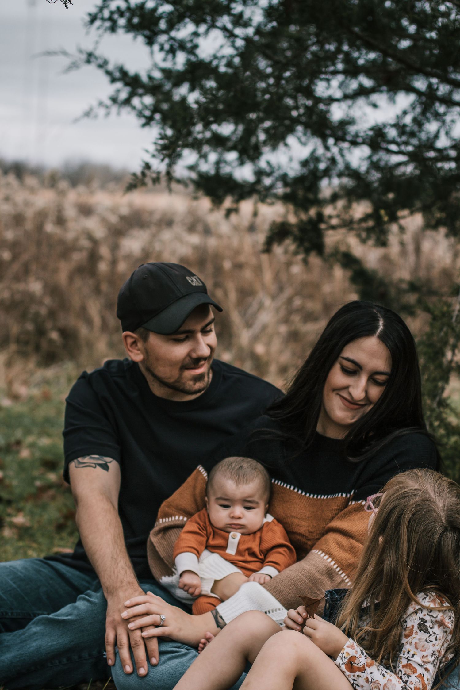 Family sits outdoors, parents smiling at their baby in a rust onesie, with a older child nearby at Oakes Quarry Park in Dayton, Ohio