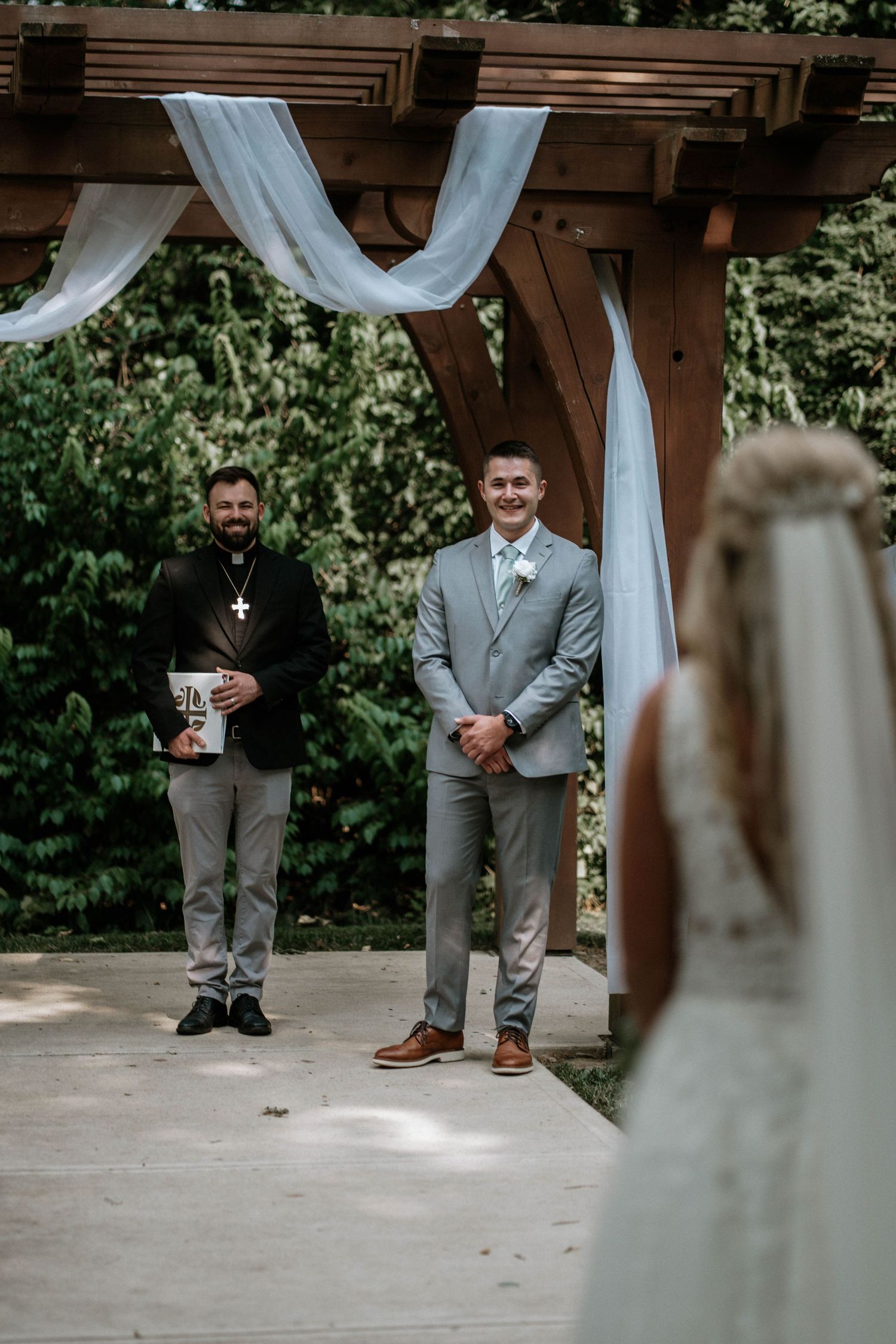 Groom in gray suit smiles at bride approaching wooden pergola altar at outdoor wedding ceremony.