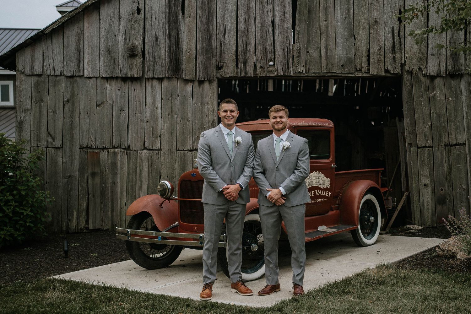 Two groomsmen in gray suits posing before a vintage red truck outside a rustic wooden barn.