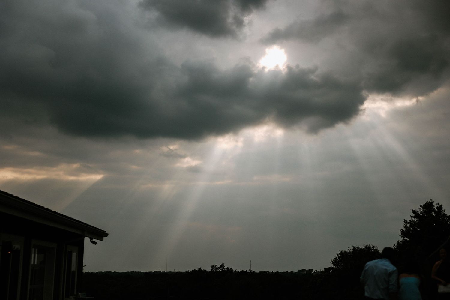 Dark storm clouds part to reveal dramatic sun rays beaming through over a treeline and building silhouette.