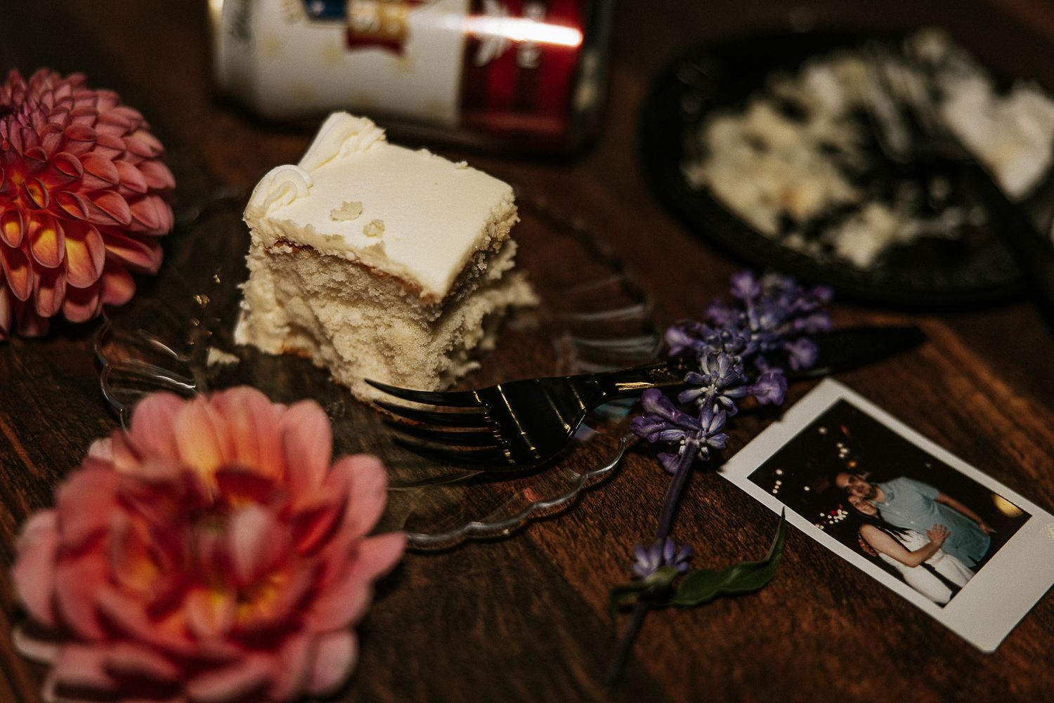 Slice of layered cake on forks beside pink dahlia, purple flowers, and a Polaroid photo on rustic wood table.