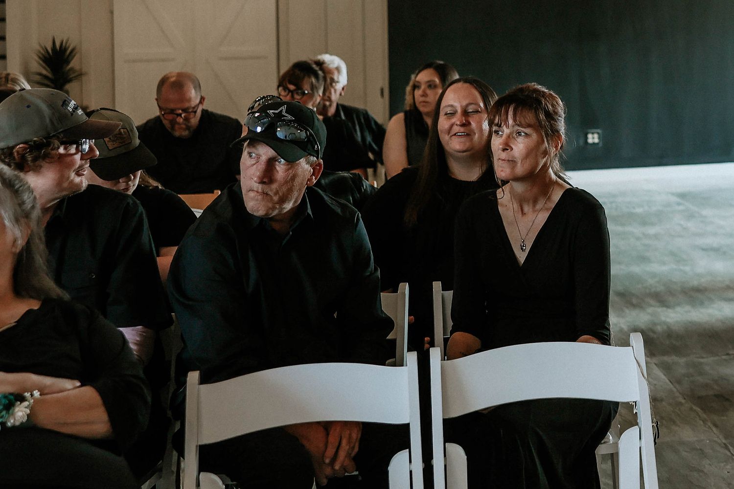 Guests in black attire seated on white folding chairs during an indoor ceremony, attentively watching the proceedings.