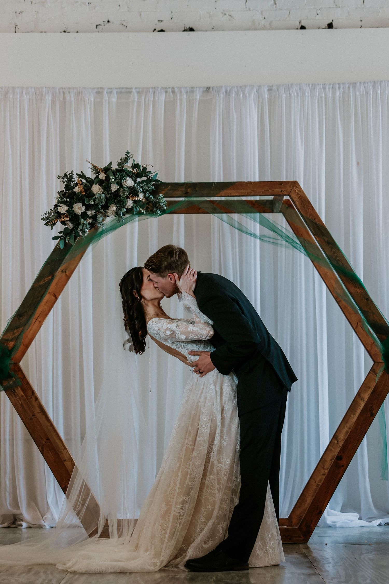 Couple shares romantic first kiss beneath floral hexagon wooden arch with white curtain backdrop.