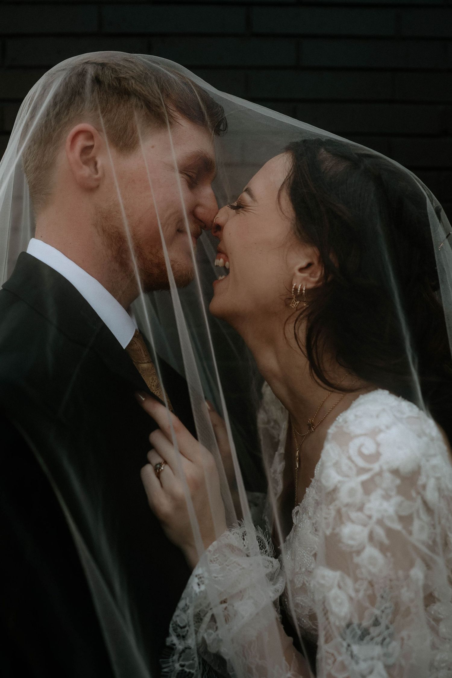 Bride and groom share an intimate moment under a wedding veil, nearly kissing, dressed in lace gown and dark suit.