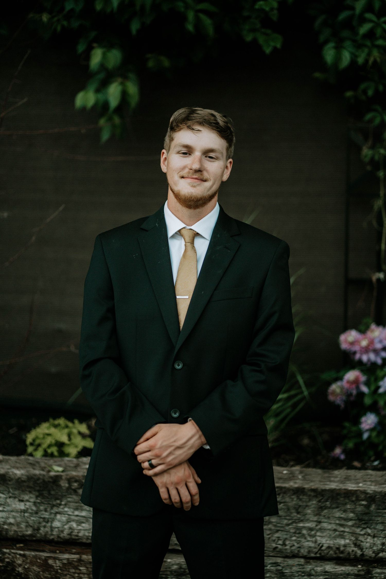 Young man in black suit and gold tie smiling outdoors with green foliage background.