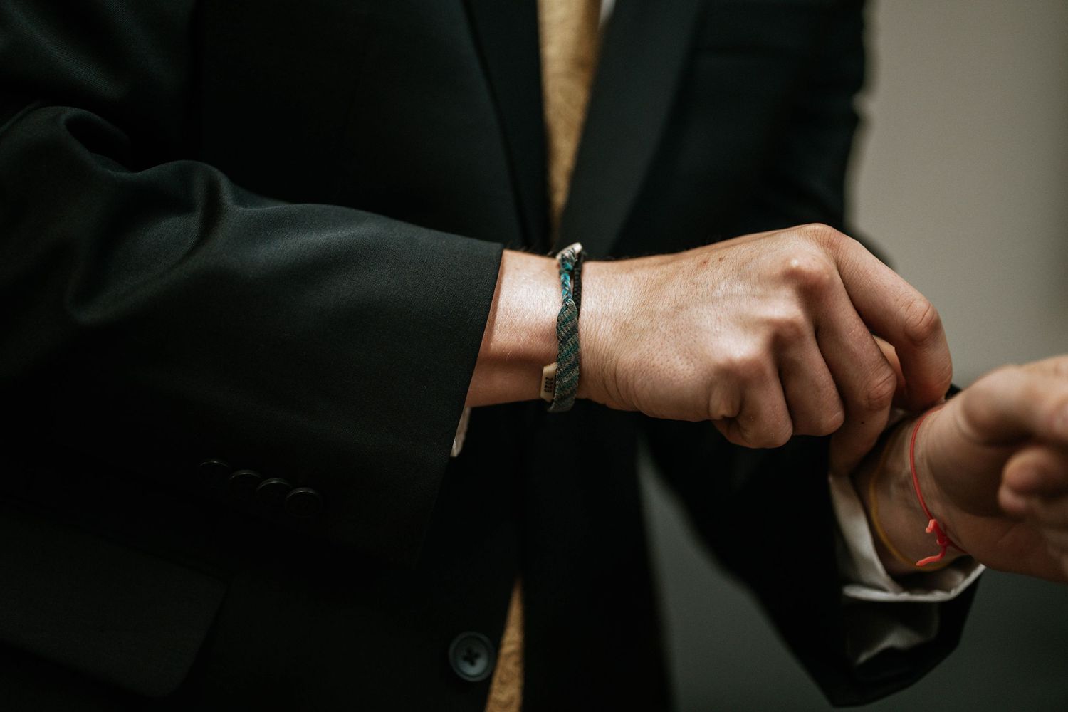 Groom in black suit adjusting cufflinks on white dress shirt with braided leather bracelet.