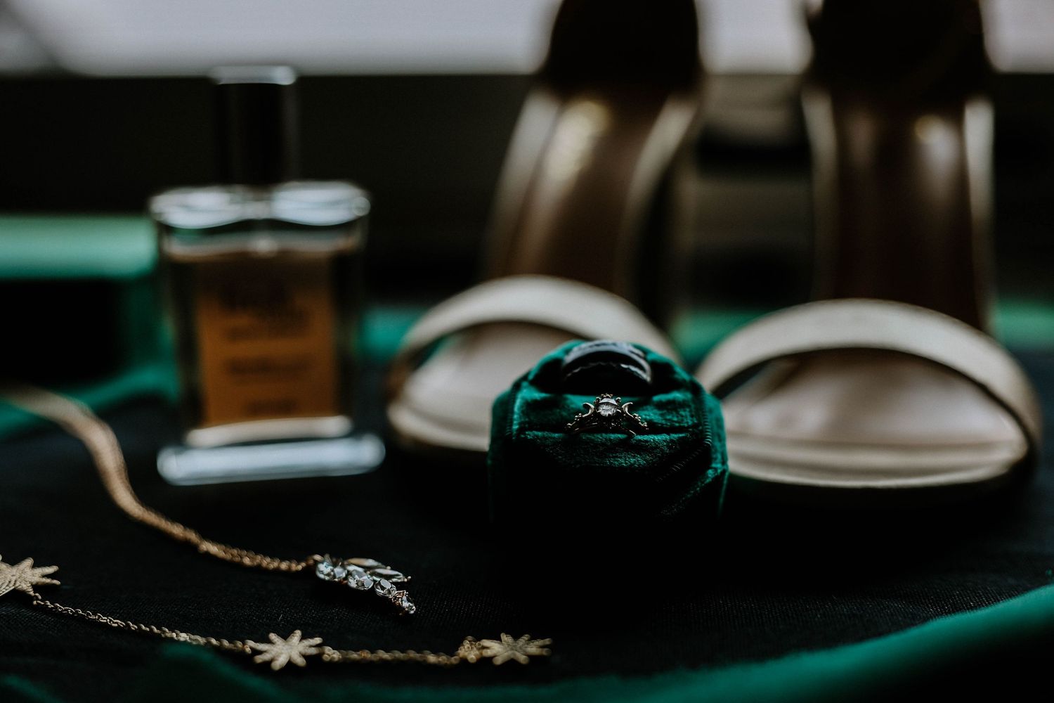 Green velvet ring box with wedding rings beside heels, perfume, and gold jewelry on emerald fabric.