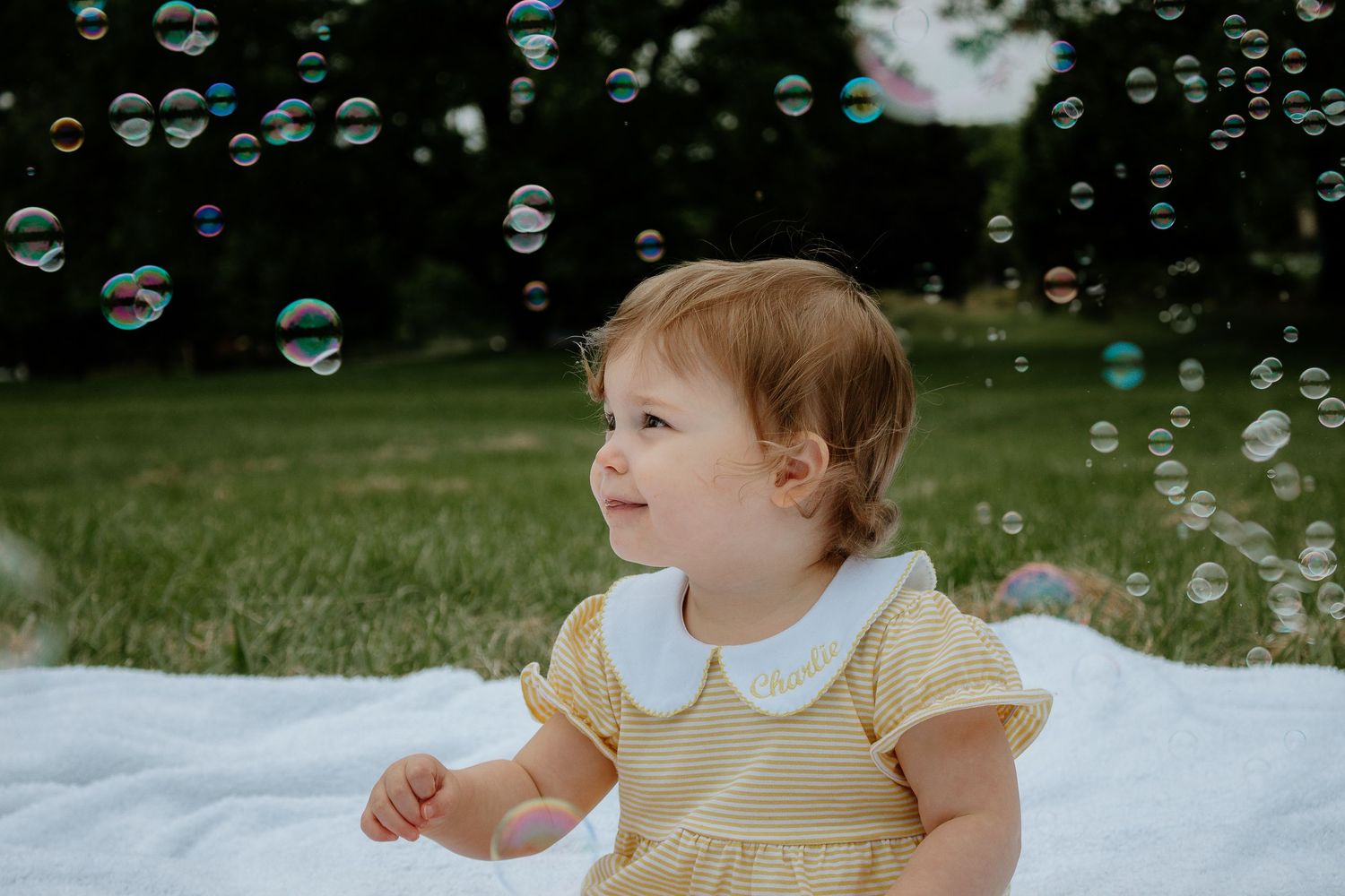 Happy baby girl in yellow dress sitting on white blanket surrounded by colorful bubbles at French Park in Cincinnati, Ohio