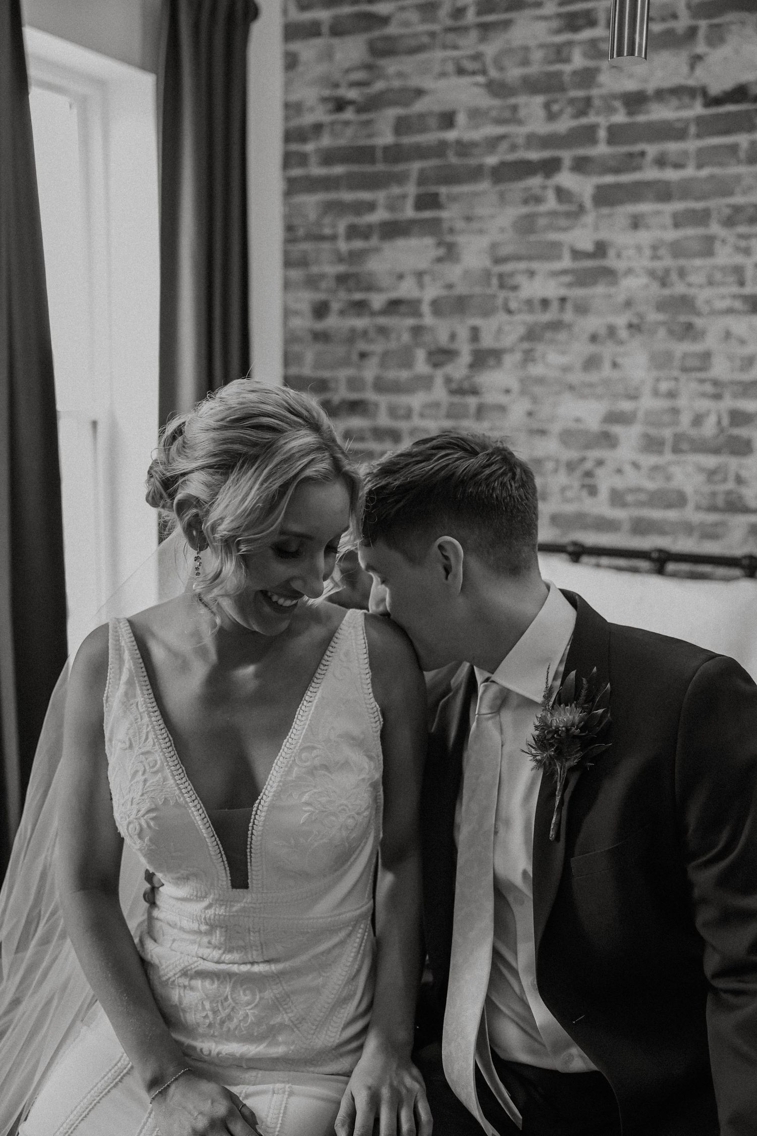Black and white photo of groom whispering to laughing bride seated before exposed brick wall at The Vue in Columbus, Ohio