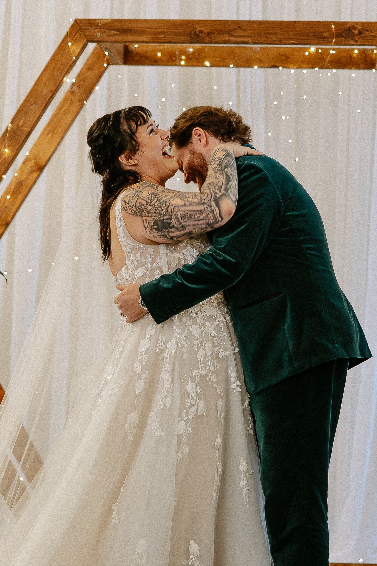 Tattooed bride and groom in green velvet suit share joyful first kiss at altar.