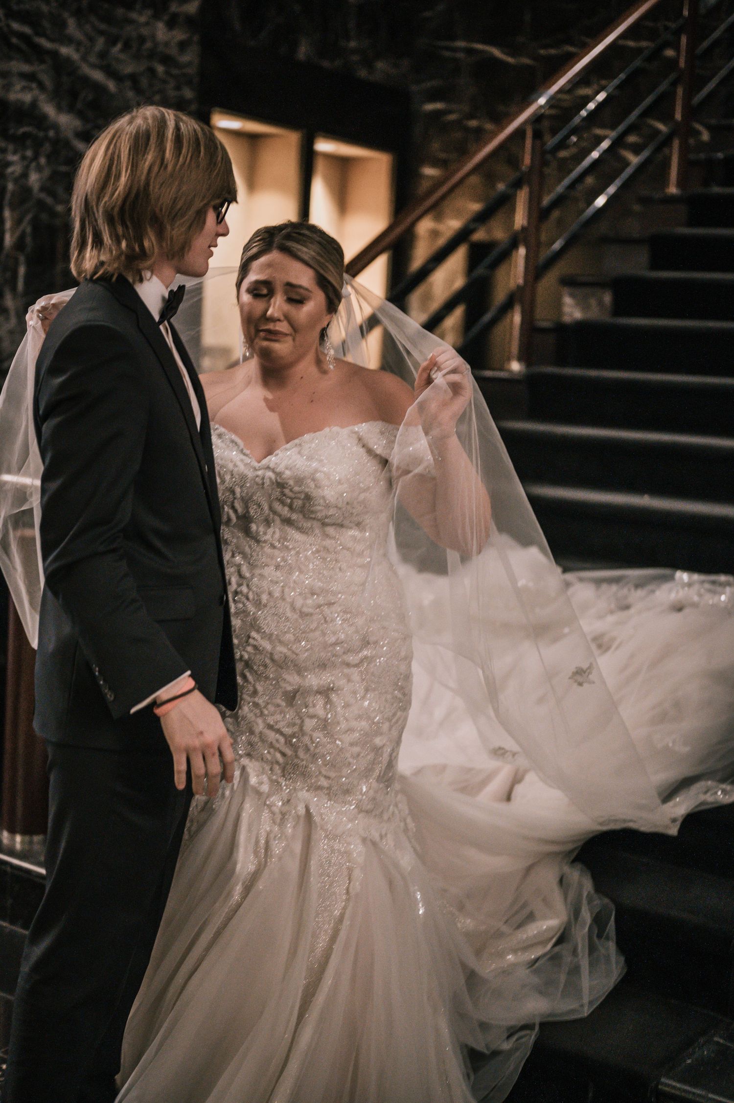 Bride in lace mermaid gown with flowing veil sharing emotional moment with groom on dark staircase.