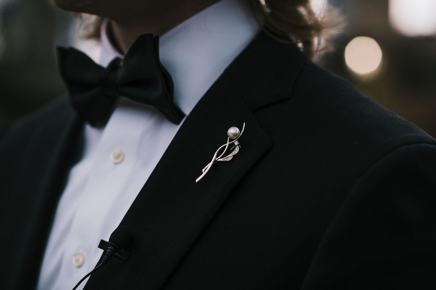 Groom's black tuxedo lapel featuring a delicate pearl and gold pin brooch with black bow tie.