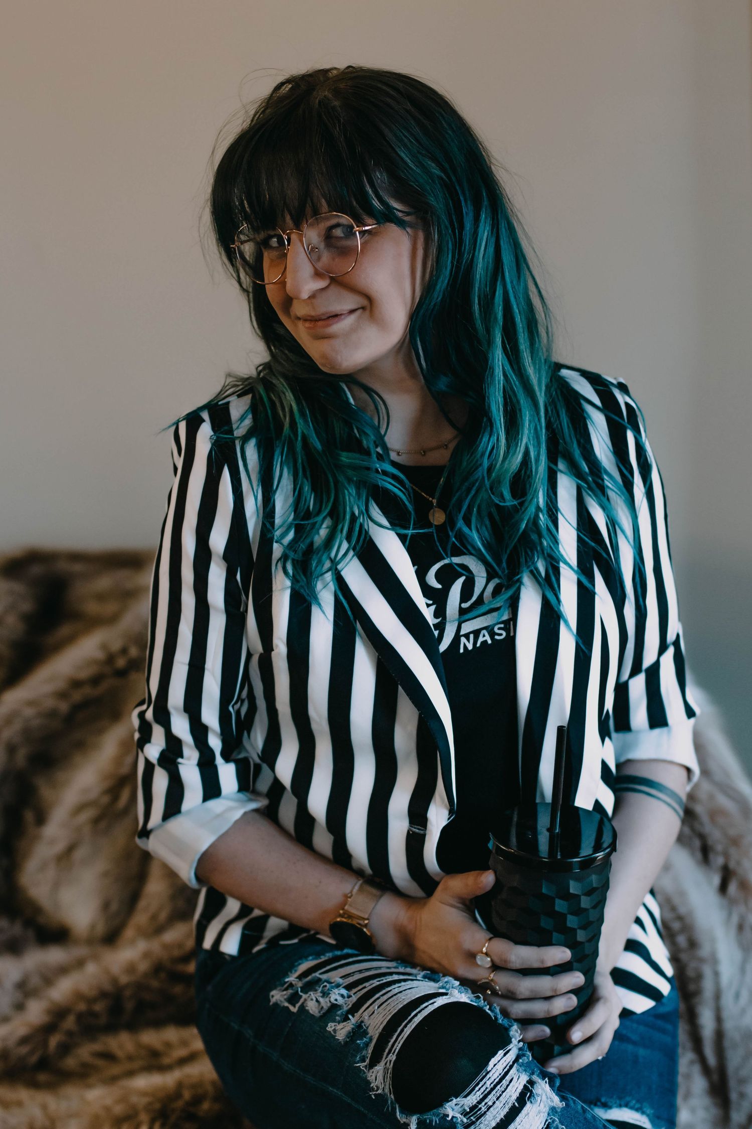 Woman with teal hair wearing striped blazer and ripped jeans smiling softly in a casual portrait.
