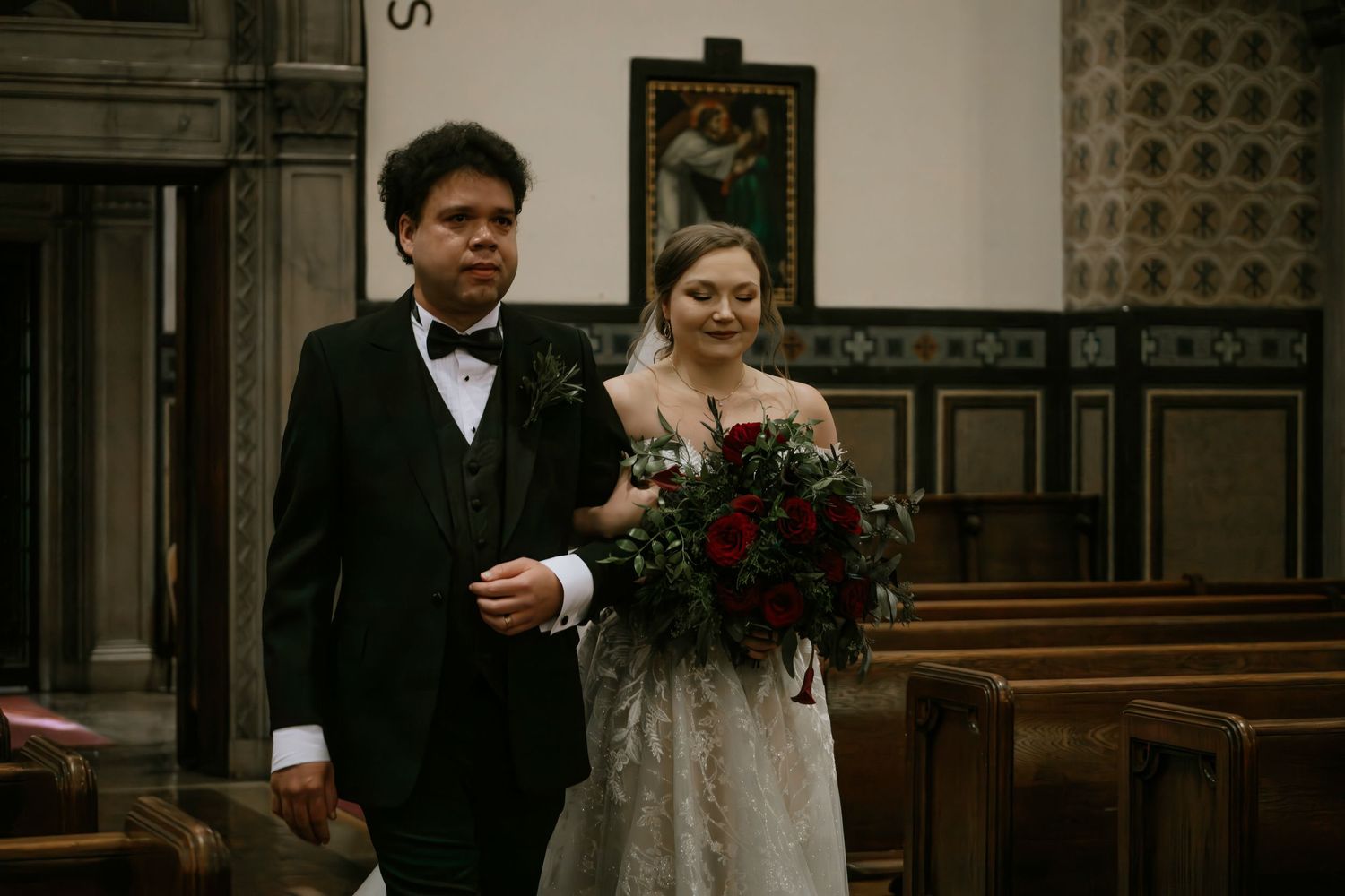 Bride in lace wedding gown carrying red bouquet walks down church aisle with escort in tuxedo in Cincinnati, Ohio