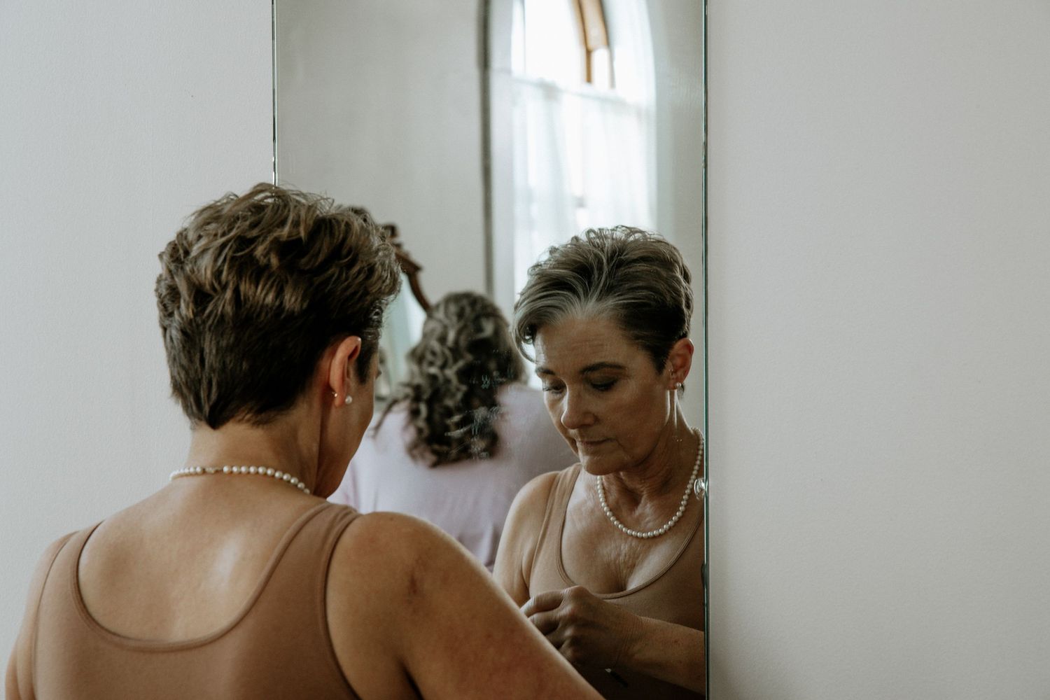 Bride wearing pearl necklace adjusting jewelry while looking in a mirror during wedding preparations at Urban Artifact in Cincinnati, Ohio