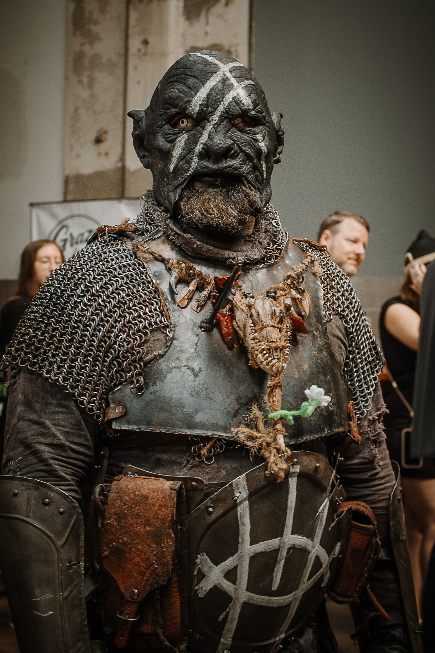 Person in detailed orc cosplay with dark face paint, chainmail armor, and weathered breastplate at a convention.