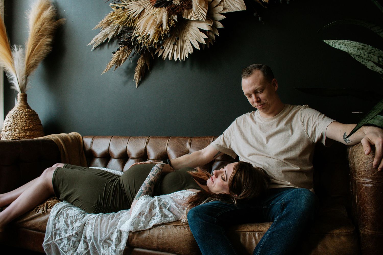 Expecting couple relaxes on a leather couch in a moody-styled room with dried floral decor at Mill Street Studios in Cincinnati, Ohio.