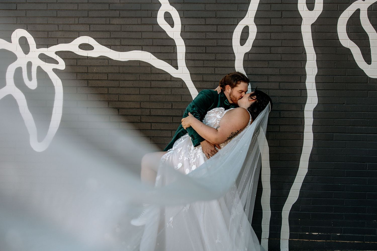 Groom dips bride in front of artistic white mural on dark brick wall, veil flowing dramatically at The Brightside in Dayton, Ohio