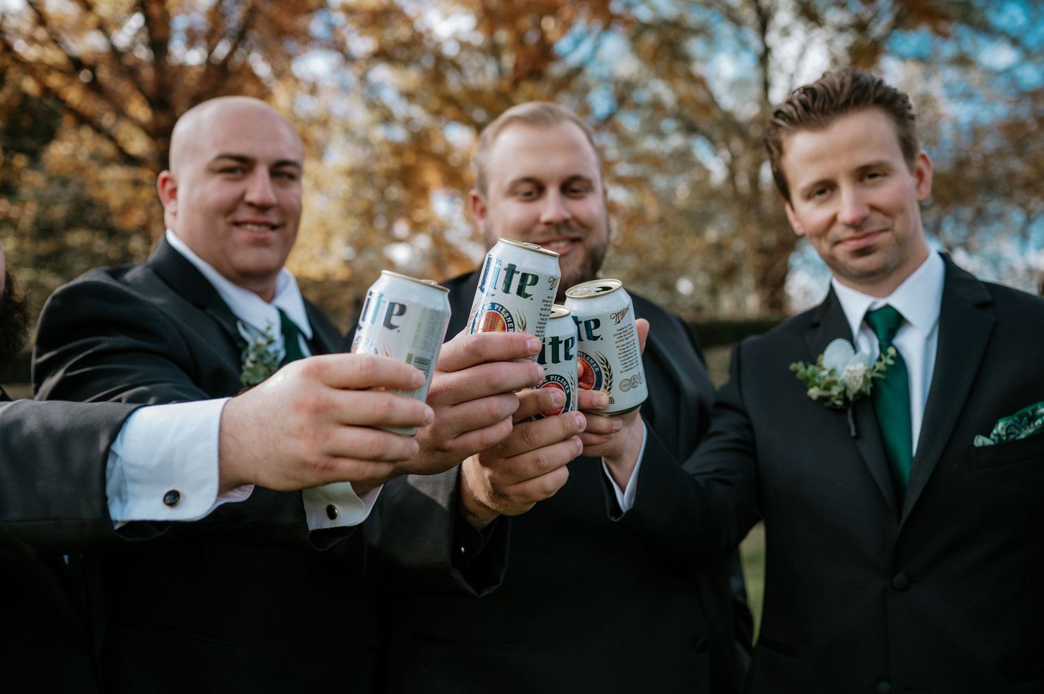 Three groomsmen in black suits toast with Miller Lite cans during a fall wedding celebration at The Red Space in Cleveland, Ohio