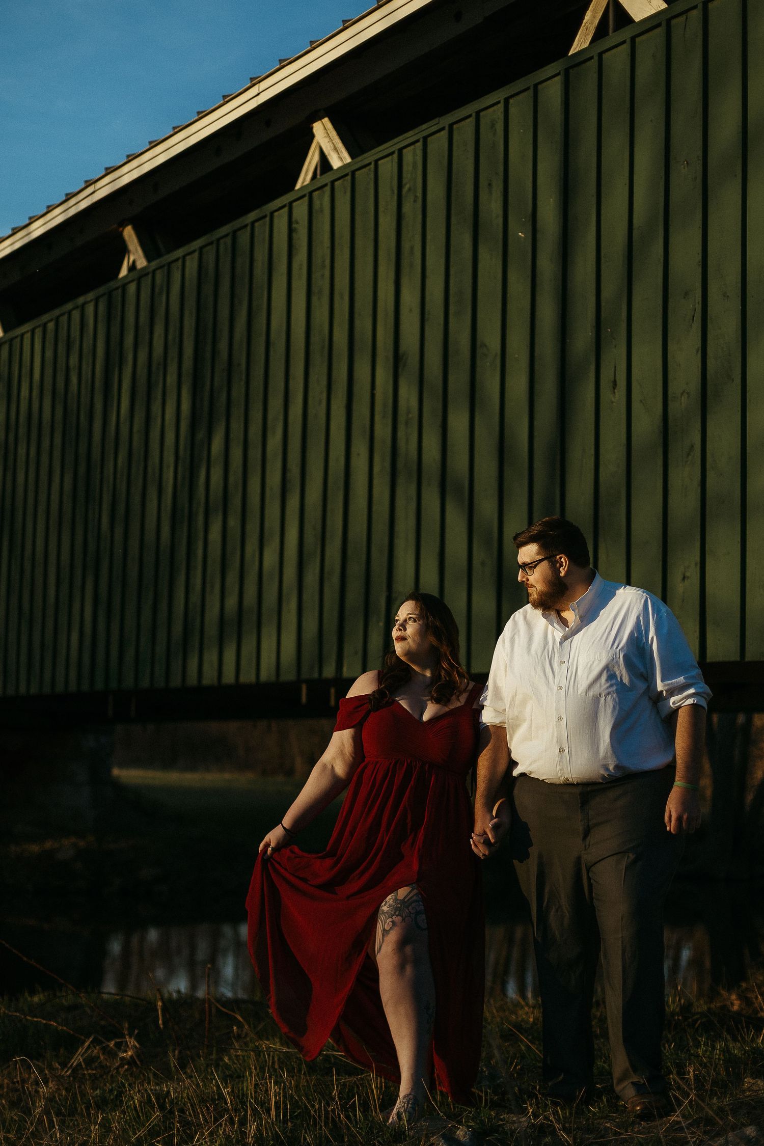 Romantic couple holding hands in golden hour light beside green barn, woman in red dress in Dayton, Ohio