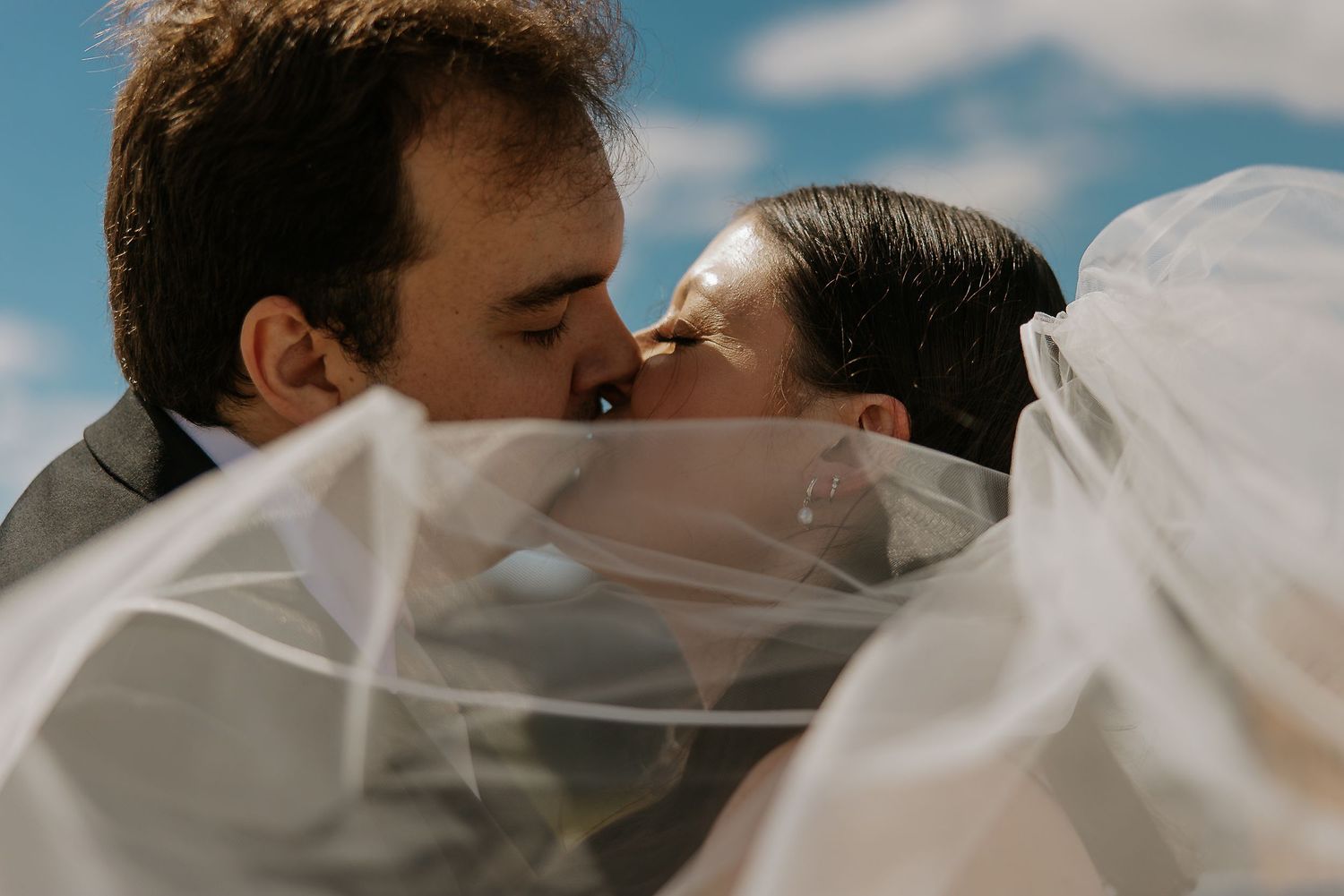 Bride and groom share a romantic kiss beneath billowing white veil against a bright blue sky.