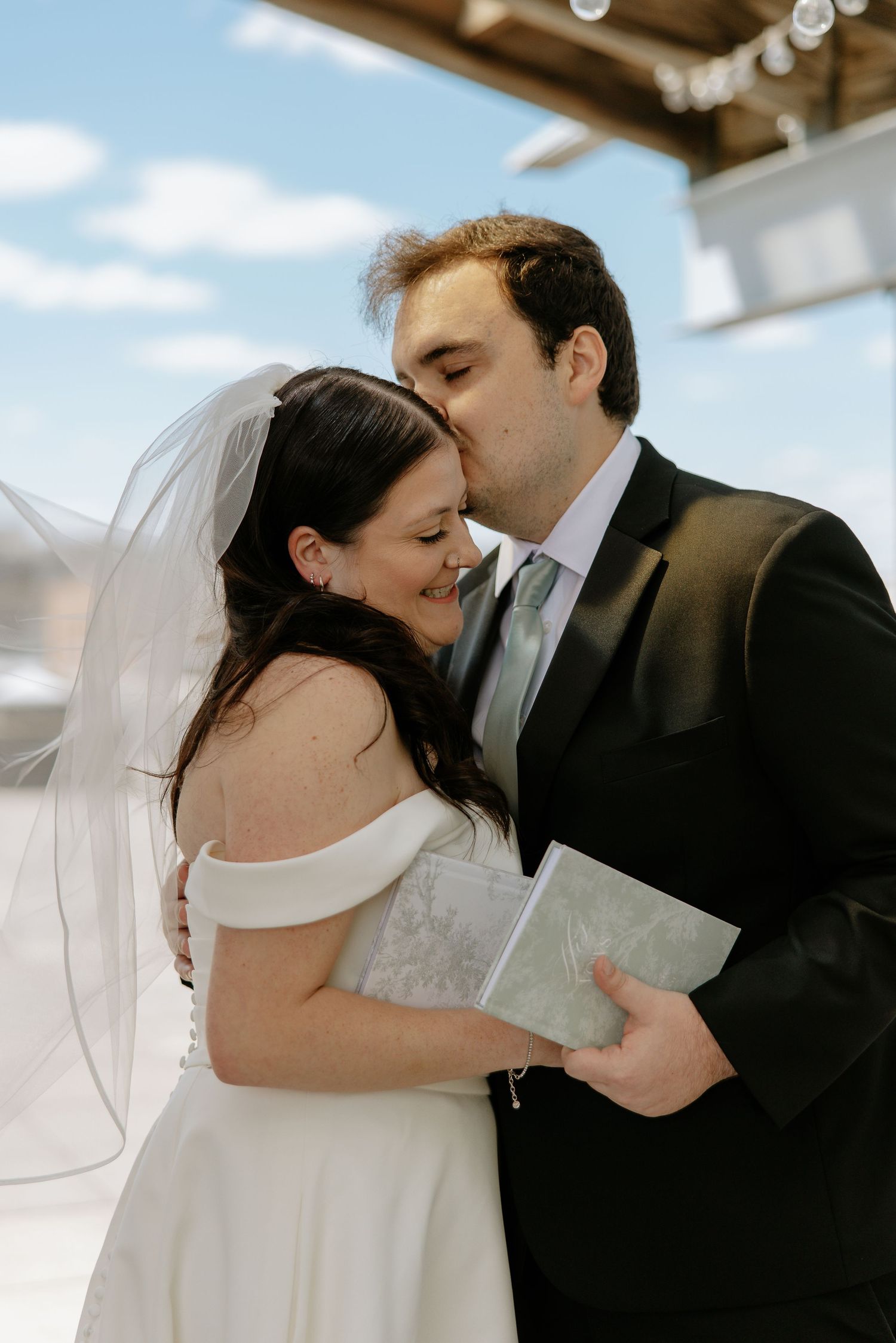 Groom kisses bride on cheek after vows, holding vow booklet, blue sky in background.