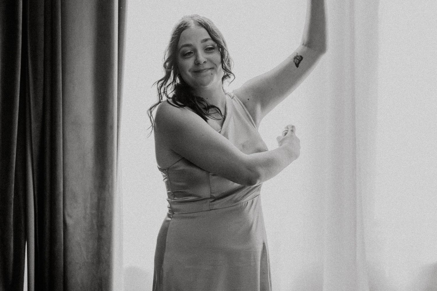 Black and white photo of smiling bridesmaid in elegant dress posing by window.