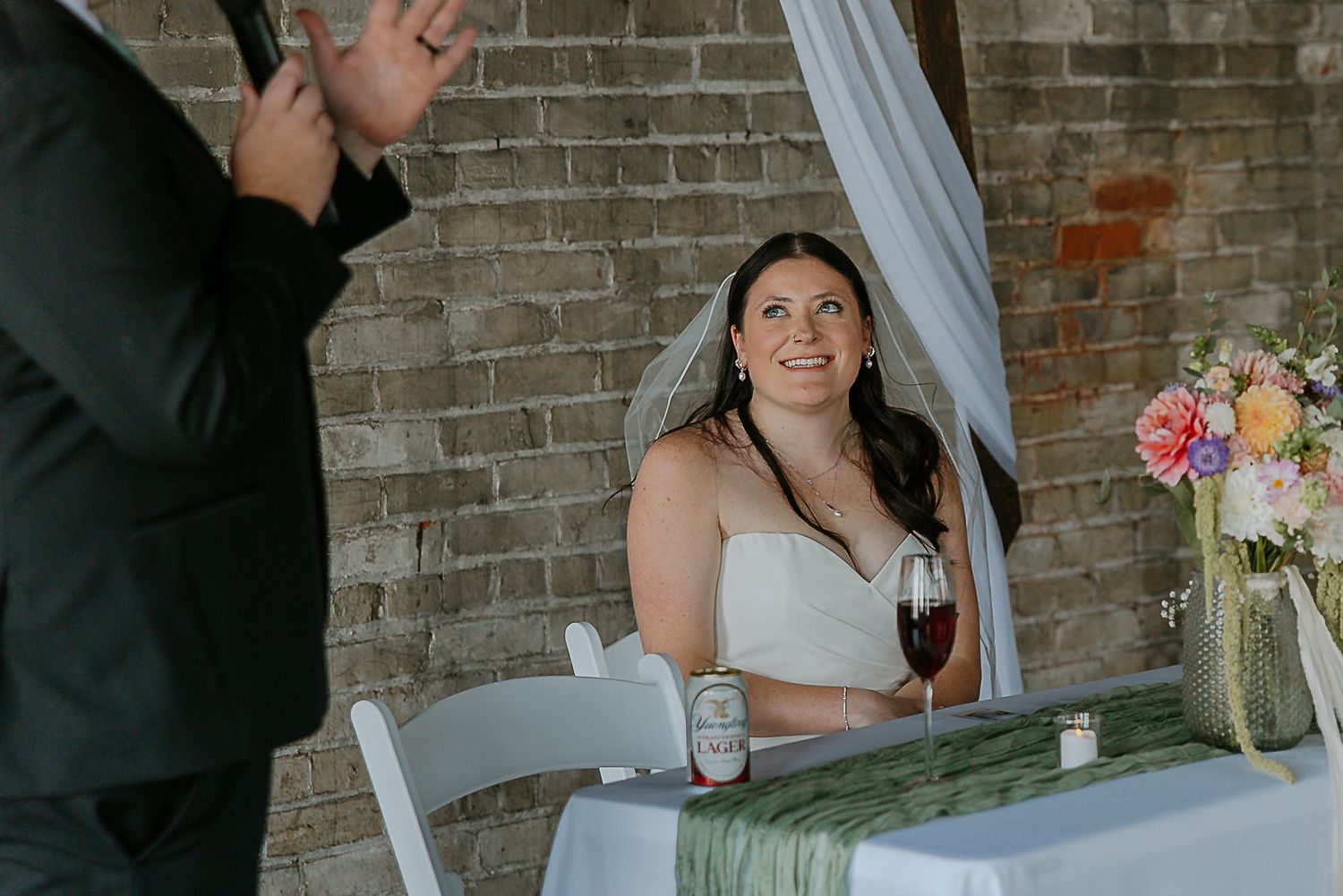 Bride holding a glass of red wine smiles up at a guest during the wedding reception.