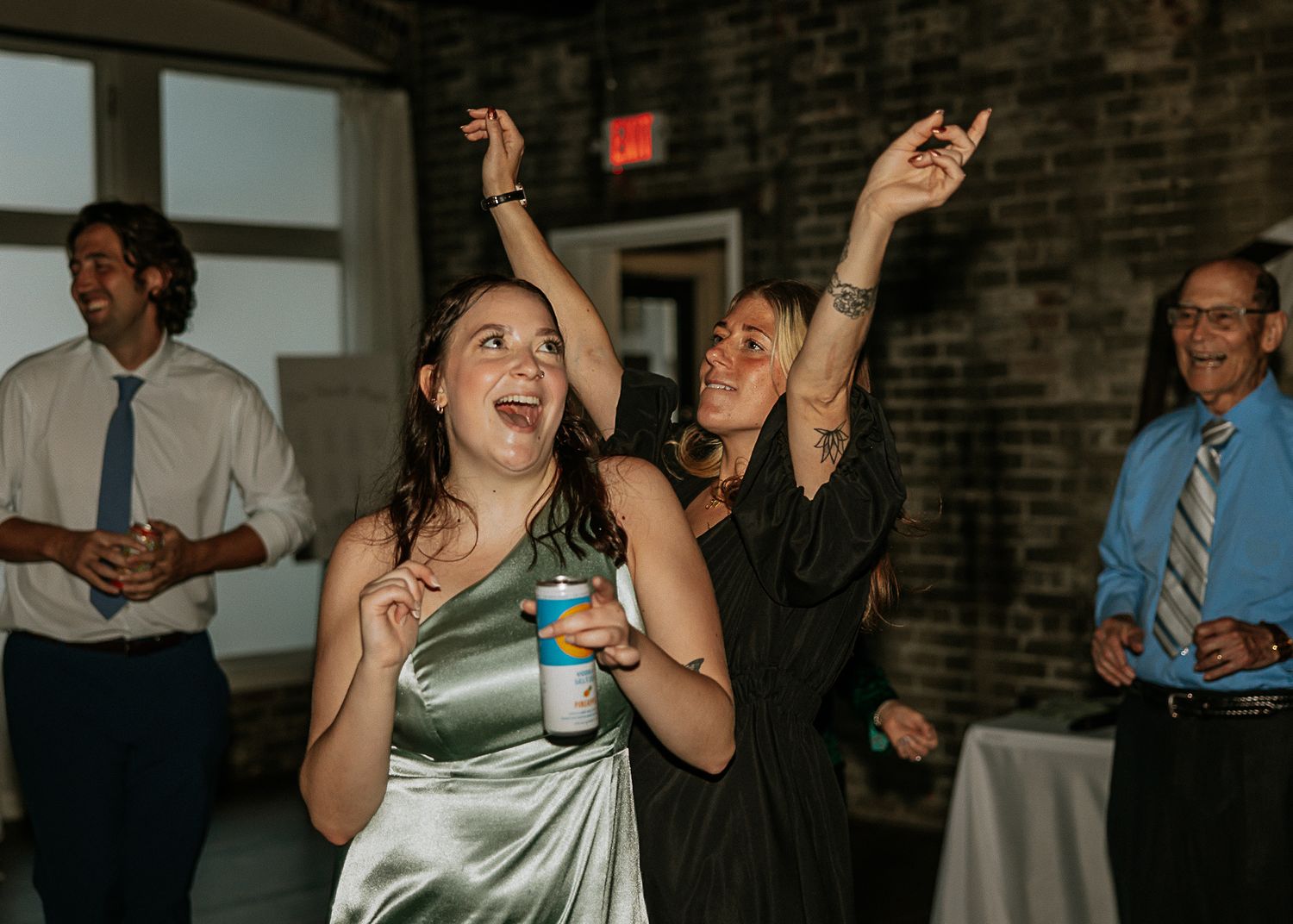 Two women in formal attire dance joyfully with arms raised at a wedding reception.