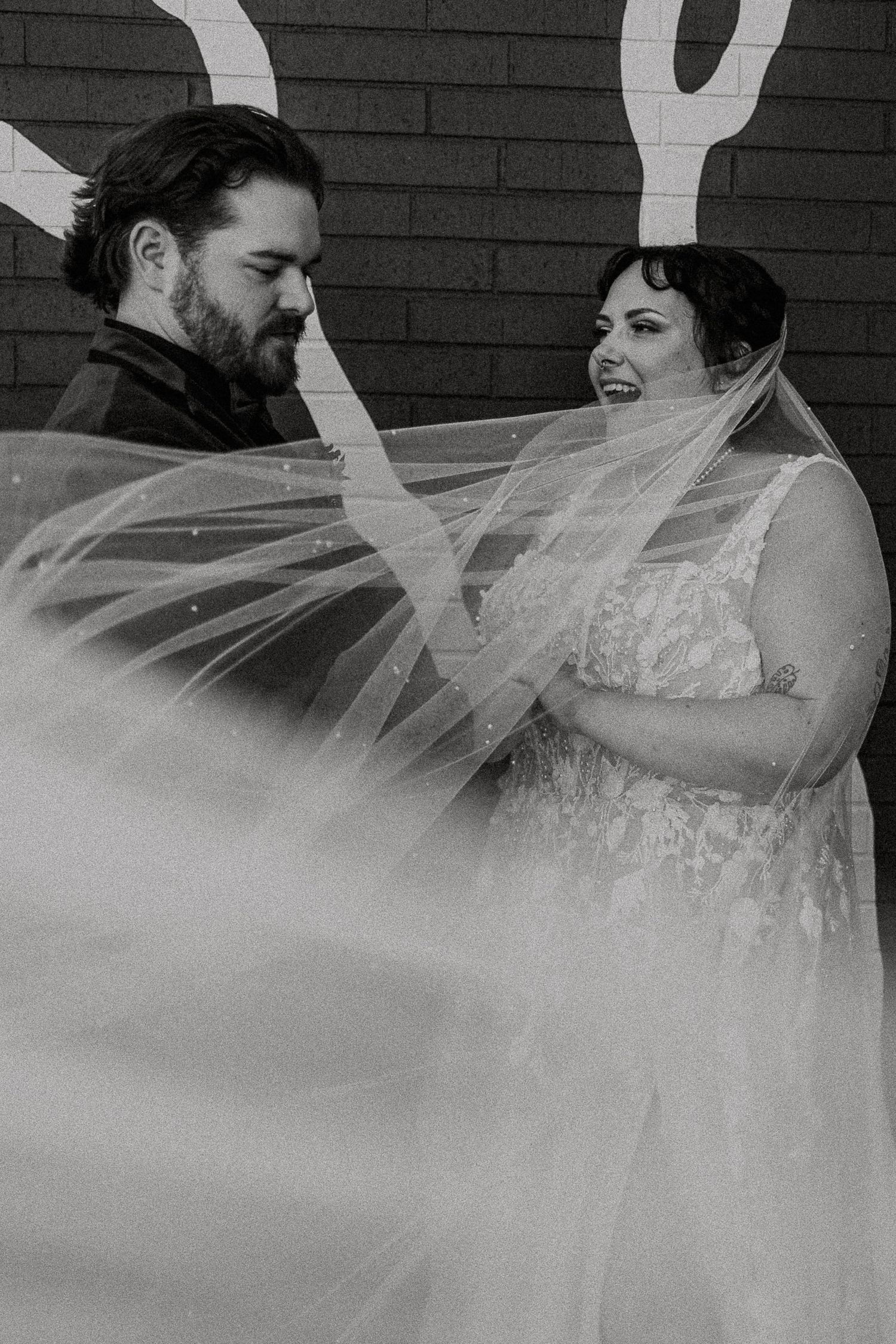 Black and white photo of laughing bride in lace dress with billowing veil beside bearded groom.
