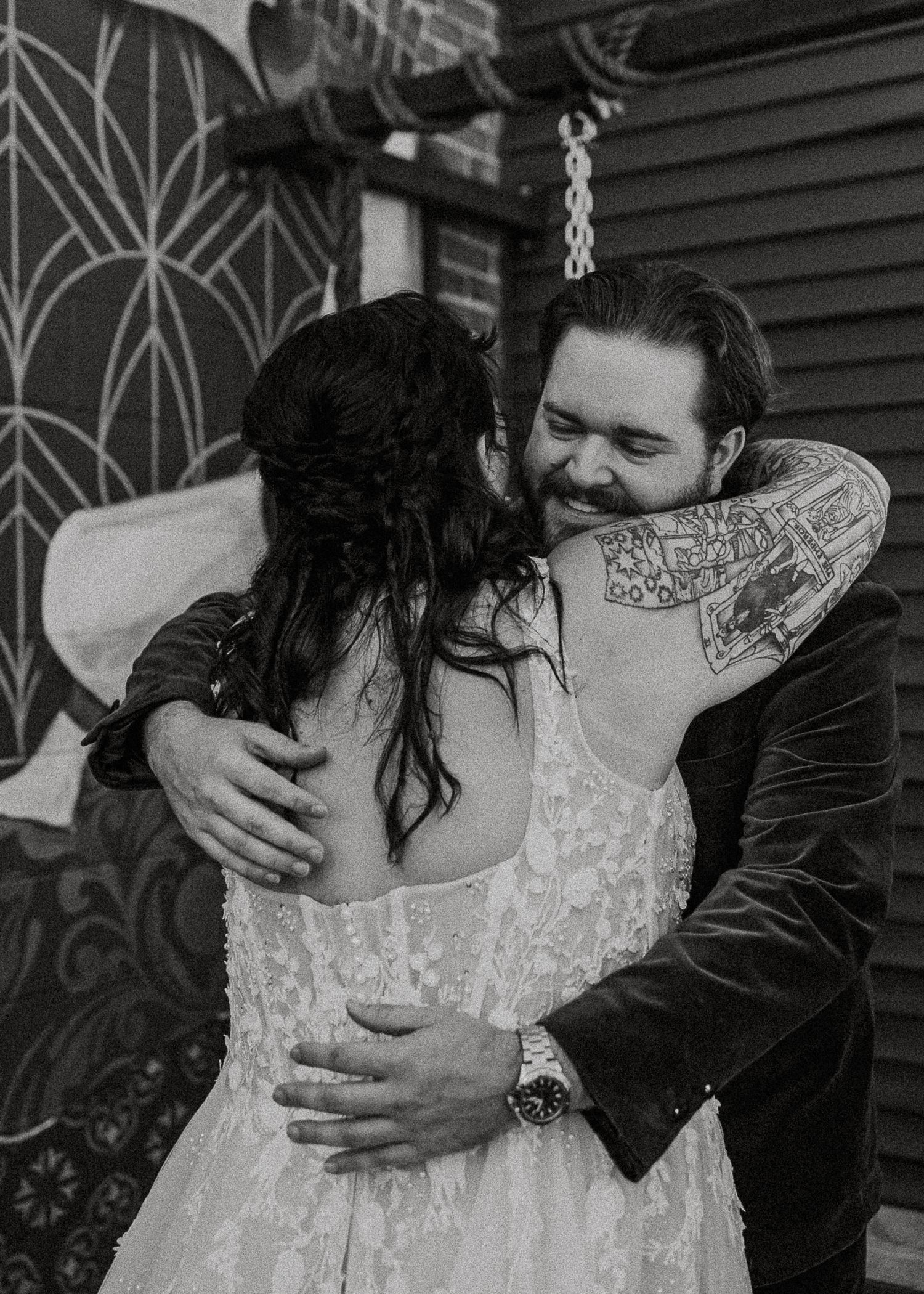 Black and white photo of groom in velvet suit embracing tattooed bride in lace wedding dress outdoors.