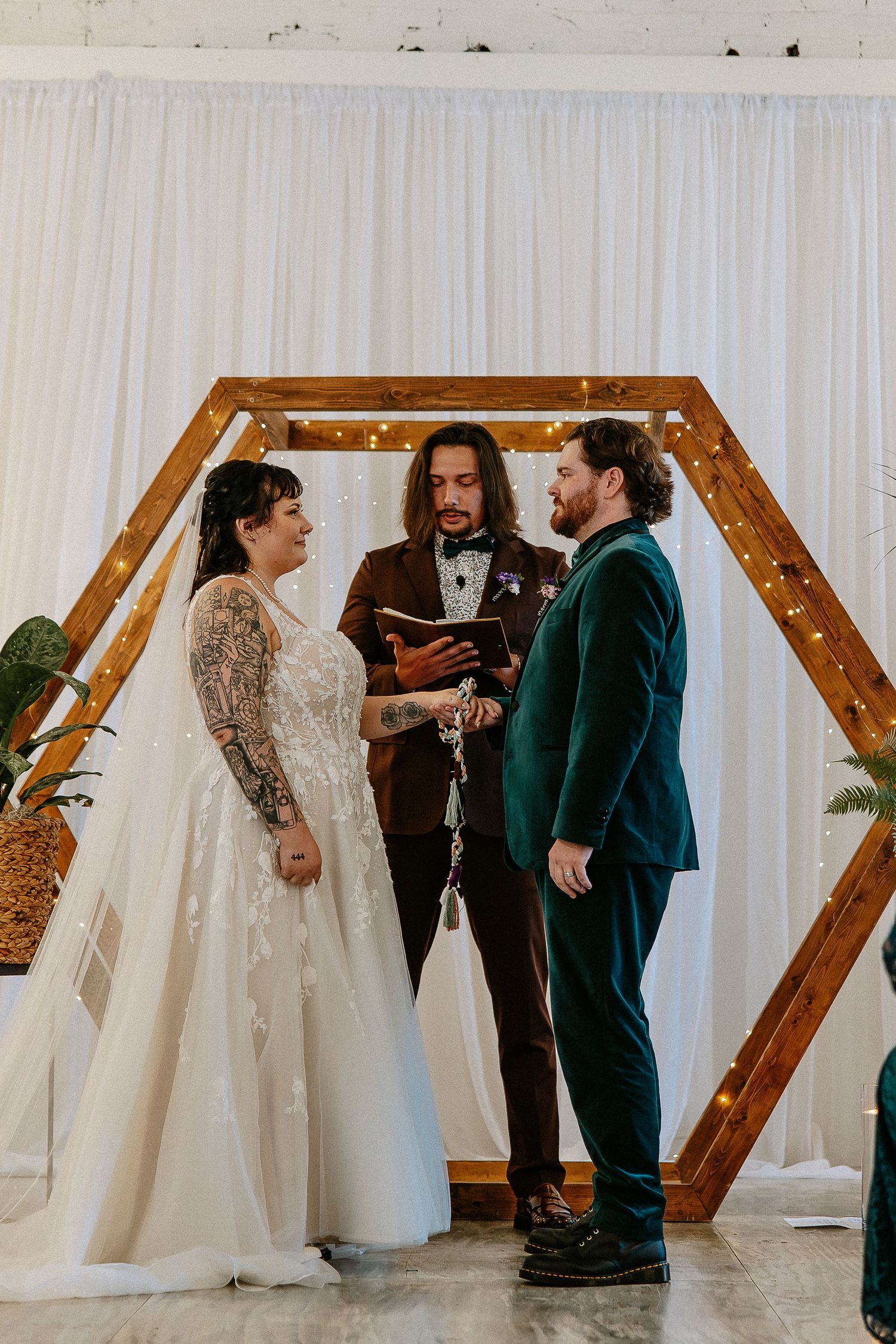 Couple exchanges vows before officiant at hexagonal wooden arch with fairy lights indoors.
