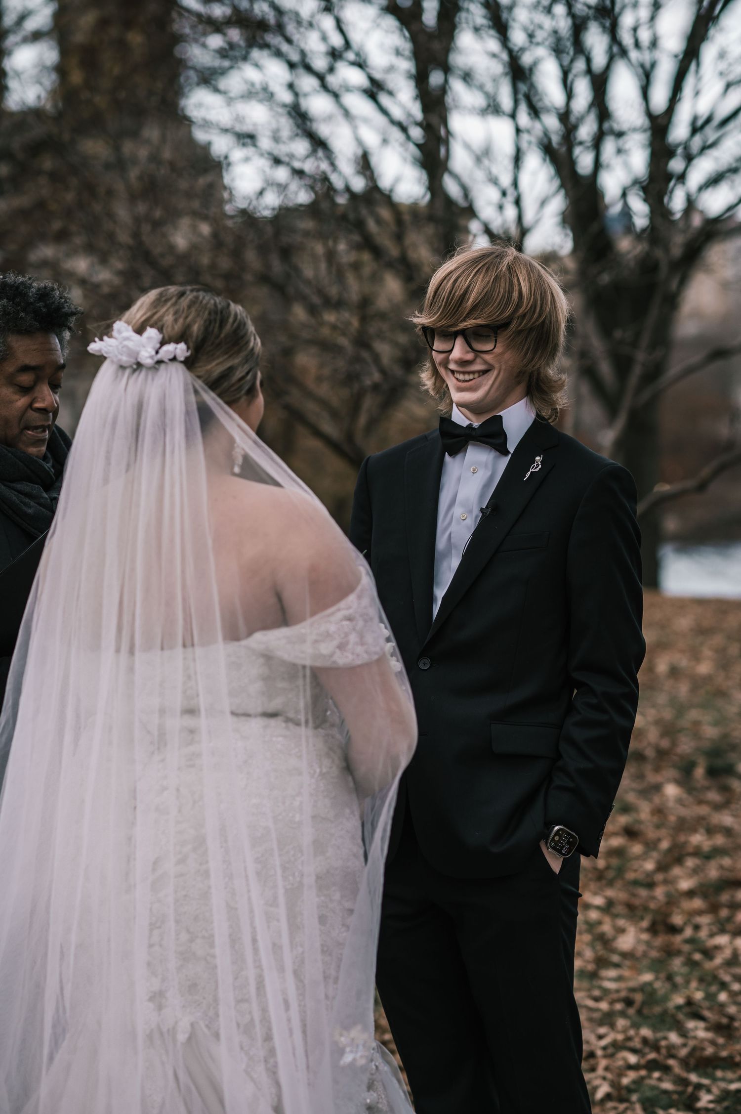 Smiling groom in black tuxedo and glasses facing veiled bride during outdoor autumn wedding ceremony.