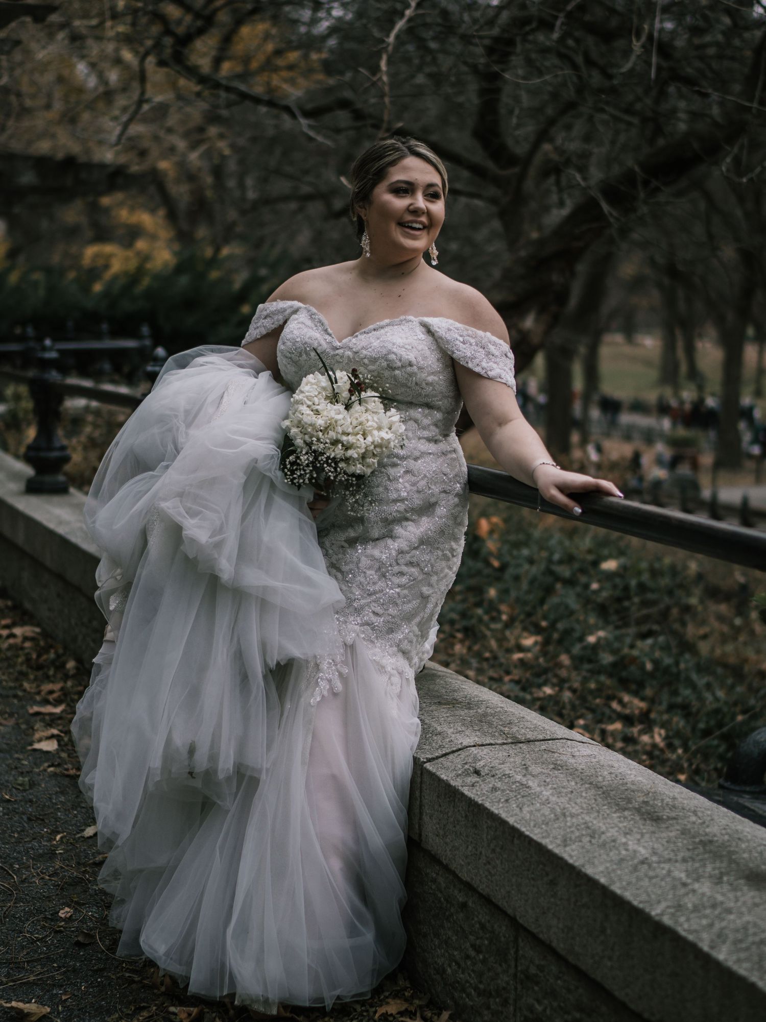Radiant bride in lace mermaid wedding dress smiles while leaning on stone wall in Central Park autumn setting.