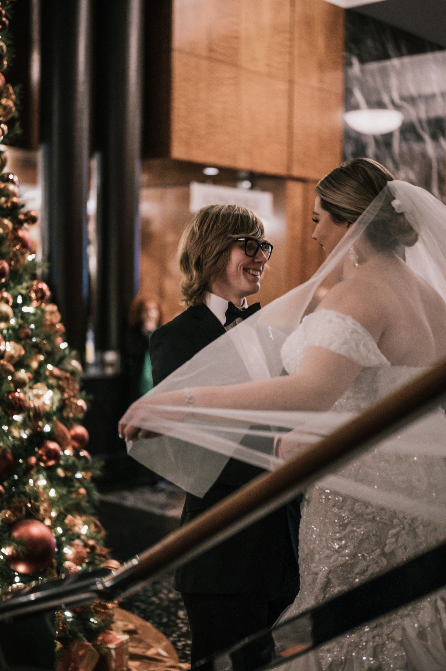 Groom in black tuxedo smiling at bride near glowing Christmas tree at wedding venue.