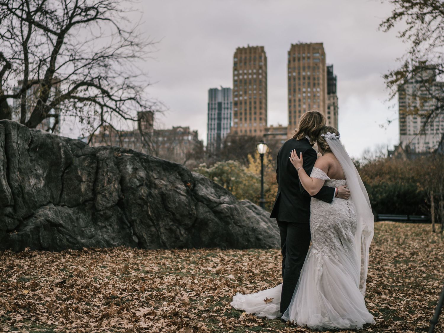 Bride and groom embrace in Central Park with New York City skyline in the background.