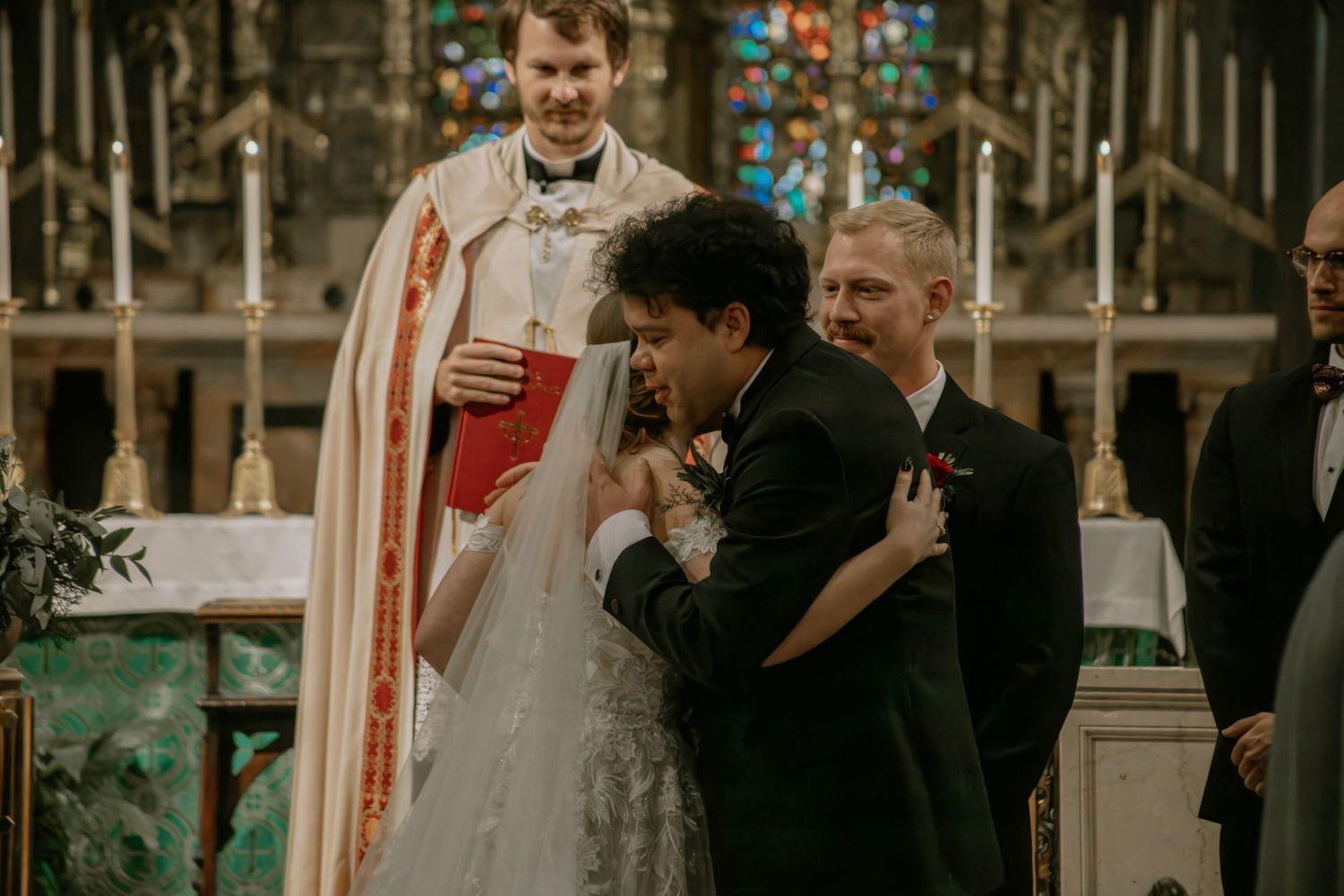 Groom emotionally embraces bride at church altar as priest and wedding party look on warmly in Cincinnati, Ohio