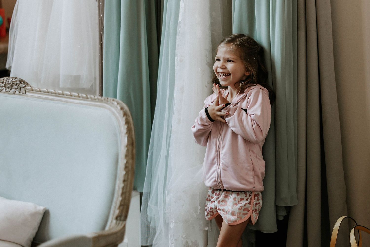 Happy little girl in pink jacket clapping with excitement surrounded by sage green wedding gowns.