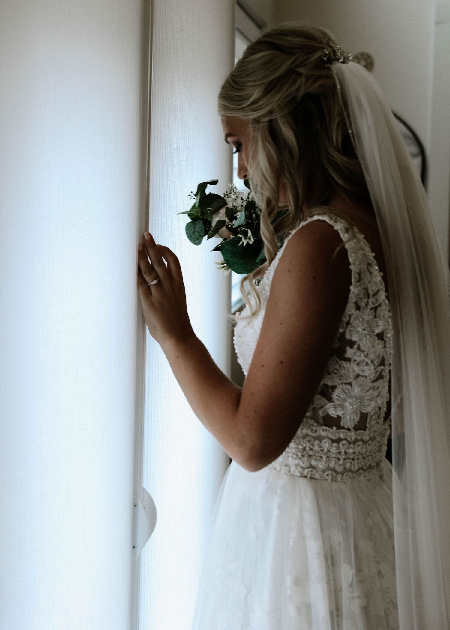 Bride in lace wedding gown with veil holding bouquet while gazing out window.