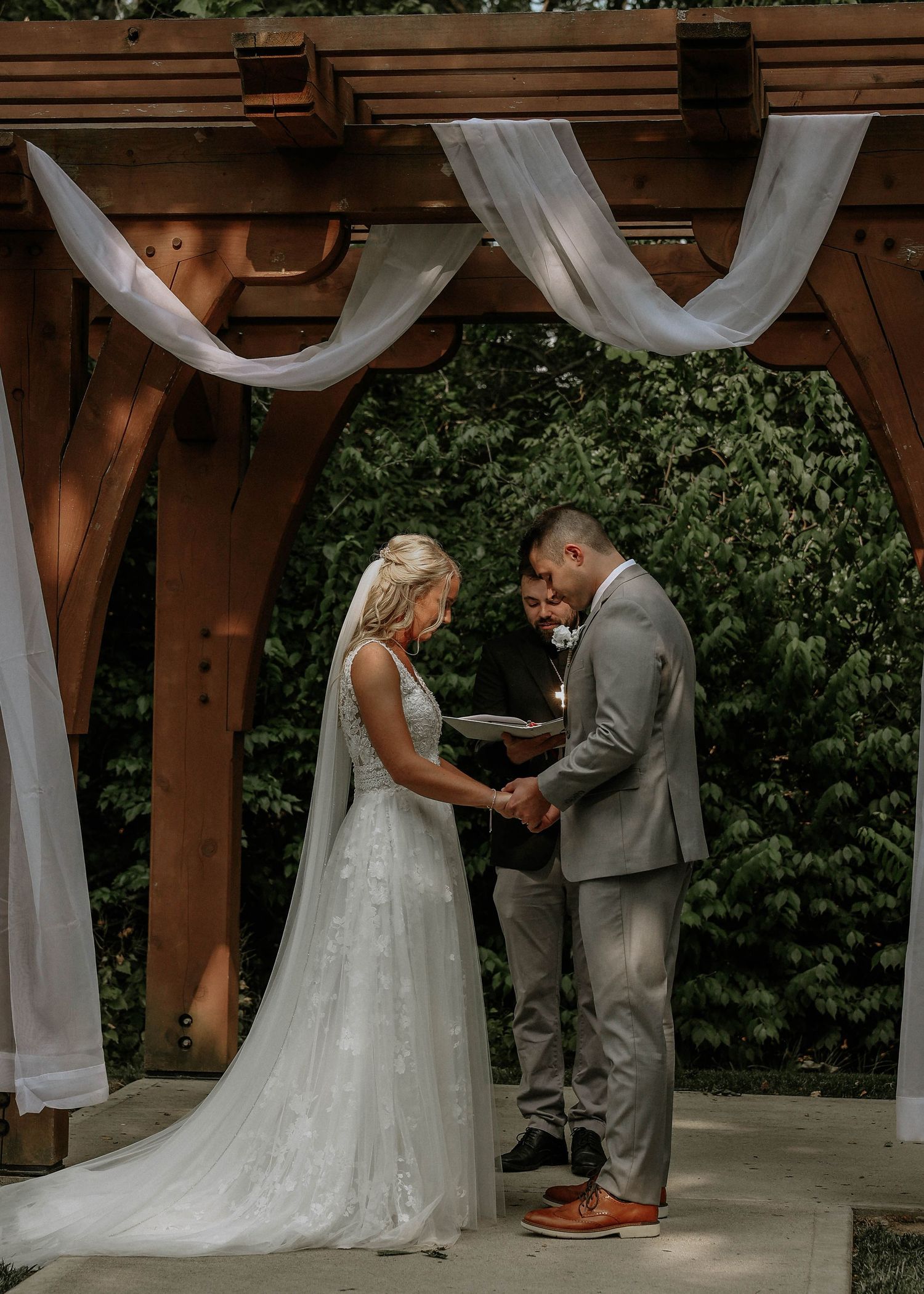 Bride and groom exchange vows under a wooden pergola draped with white fabric at an outdoor wedding ceremony.