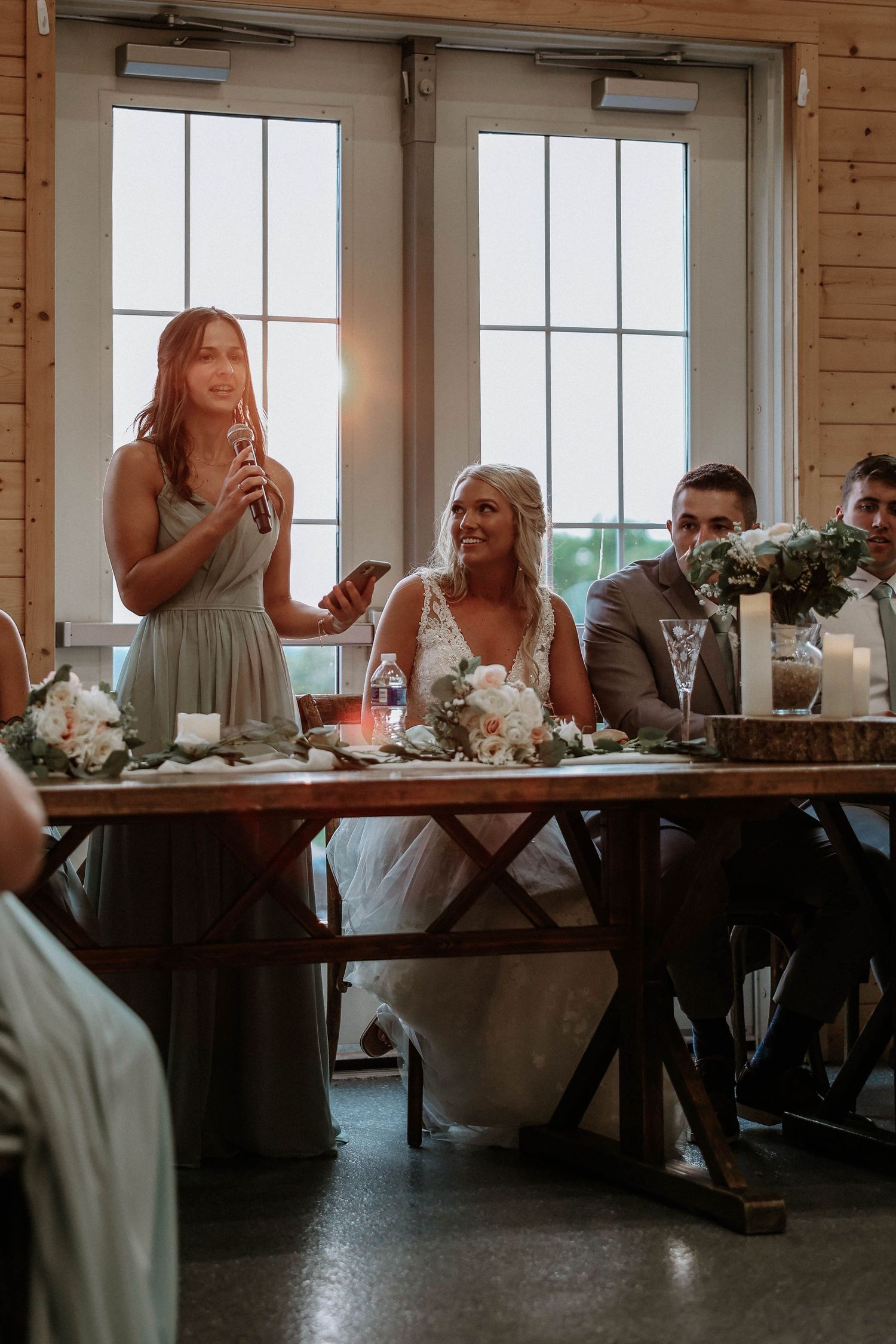 Bridesmaid giving a heartfelt speech at a rustic wedding reception while the bride smiles joyfully at the head table.