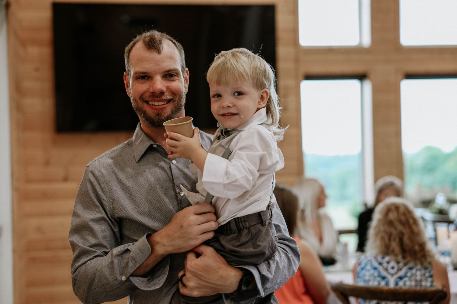 Smiling man in gray shirt holding adorable blonde toddler boy at a wedding reception venue.