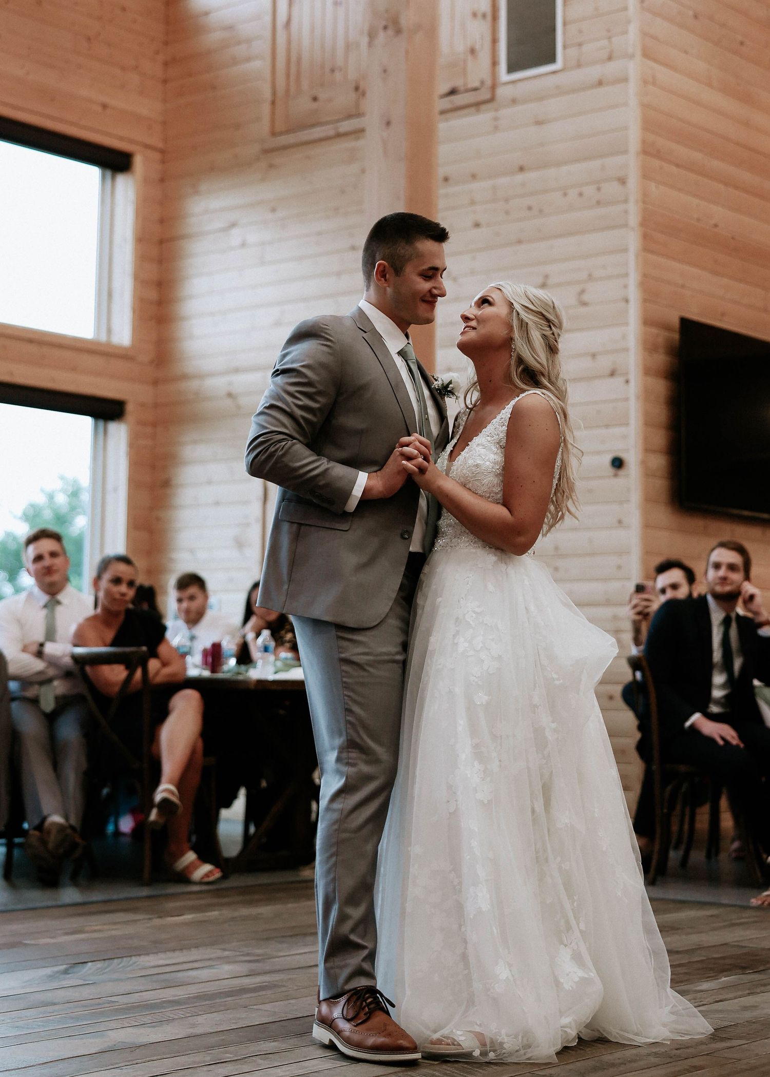 Bride and groom share their first dance in a rustic wood-paneled venue with guests watching in the background.