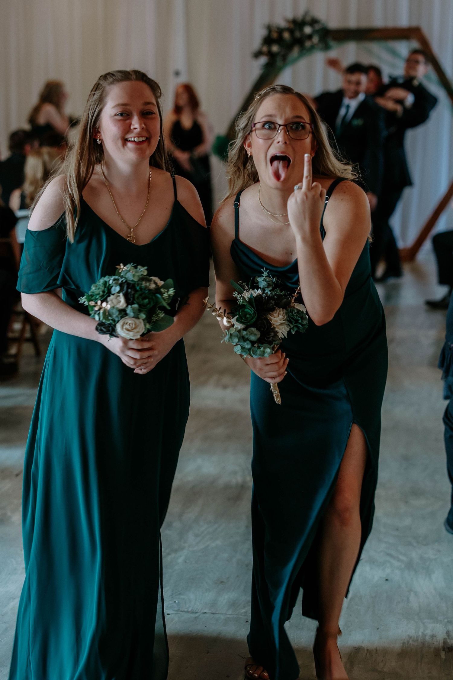 Two bridesmaids in emerald green gowns walk down the aisle holding bouquets at a wedding ceremony.