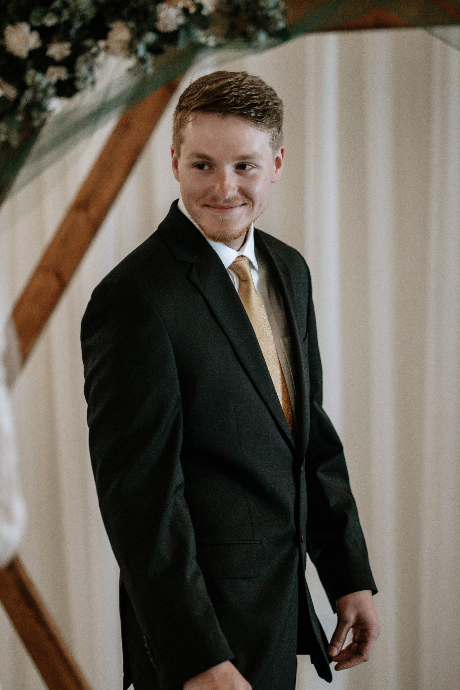 Smiling groom in black suit and gold tie standing at wedding altar with floral arch backdrop.