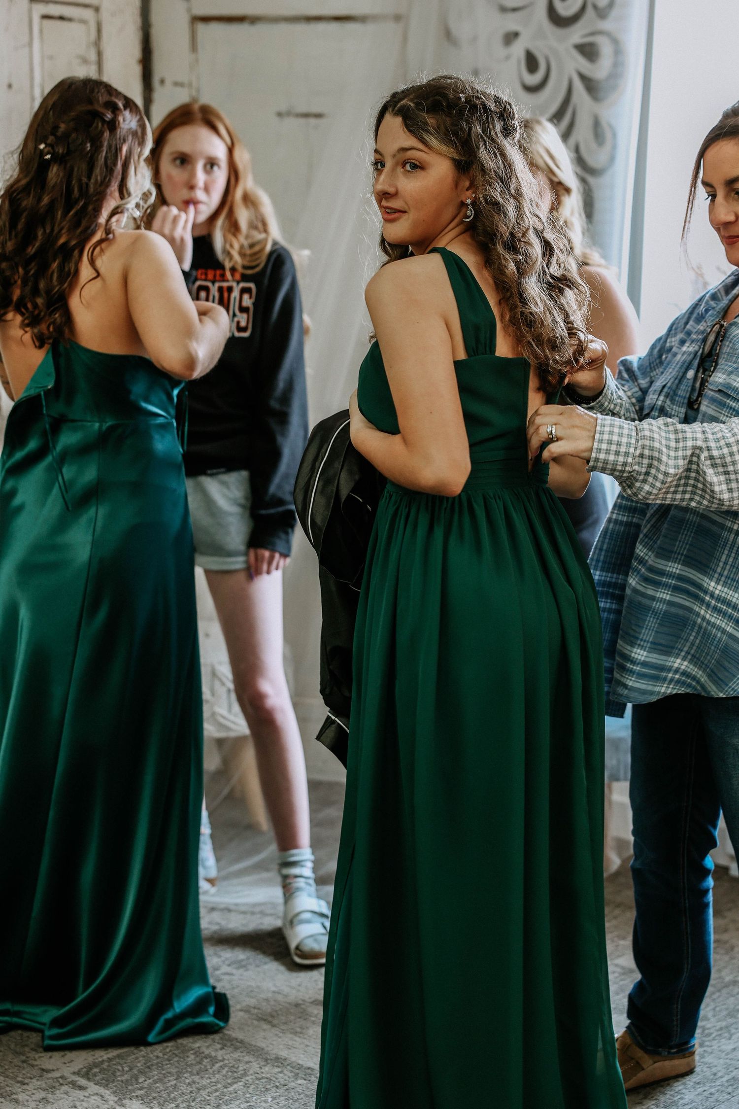 Two women in elegant emerald green satin gowns stand in a room, one glancing back over her shoulder.