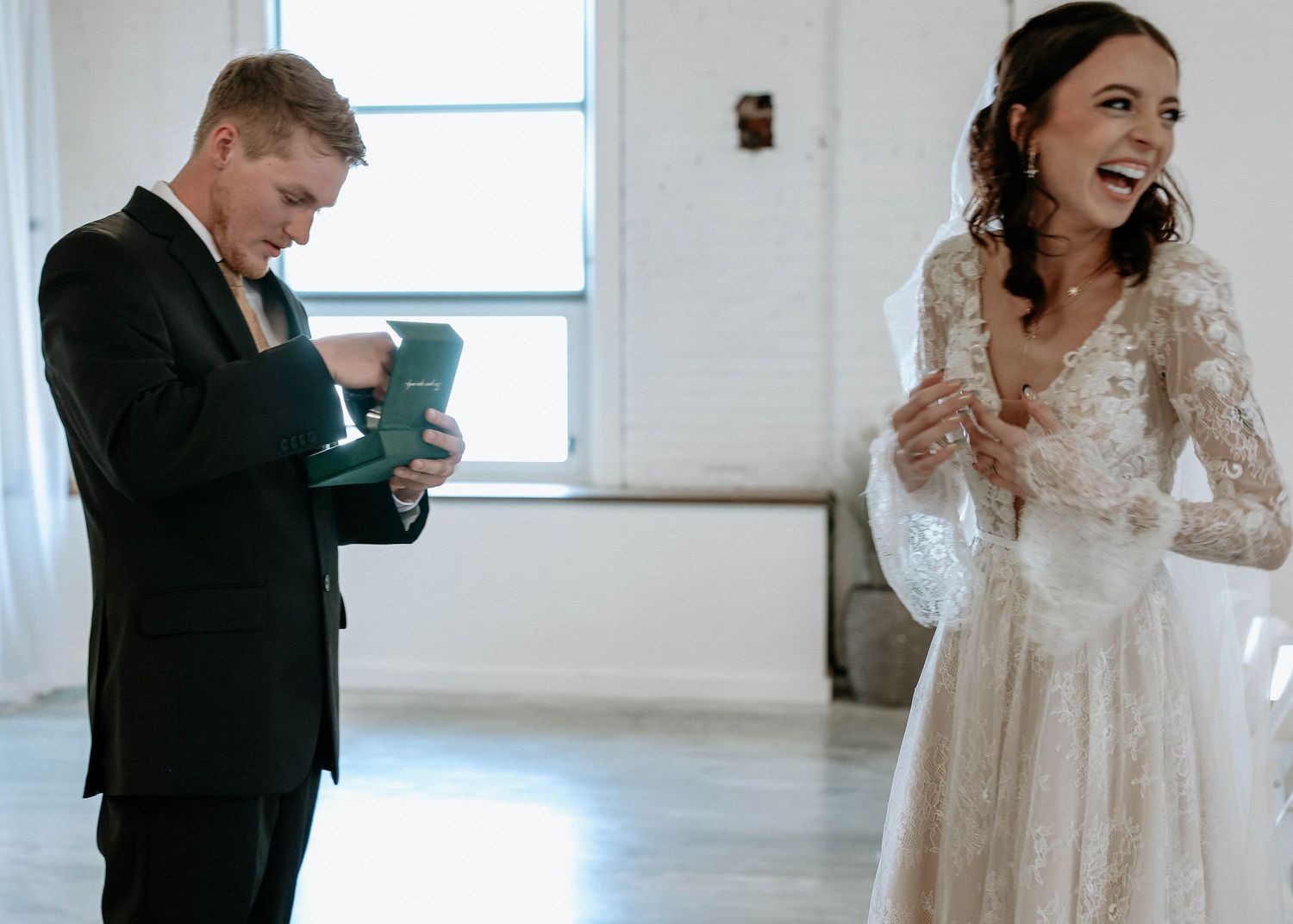 Bride laughing in lace gown as groom reads vows from teal box during emotional wedding first look.
