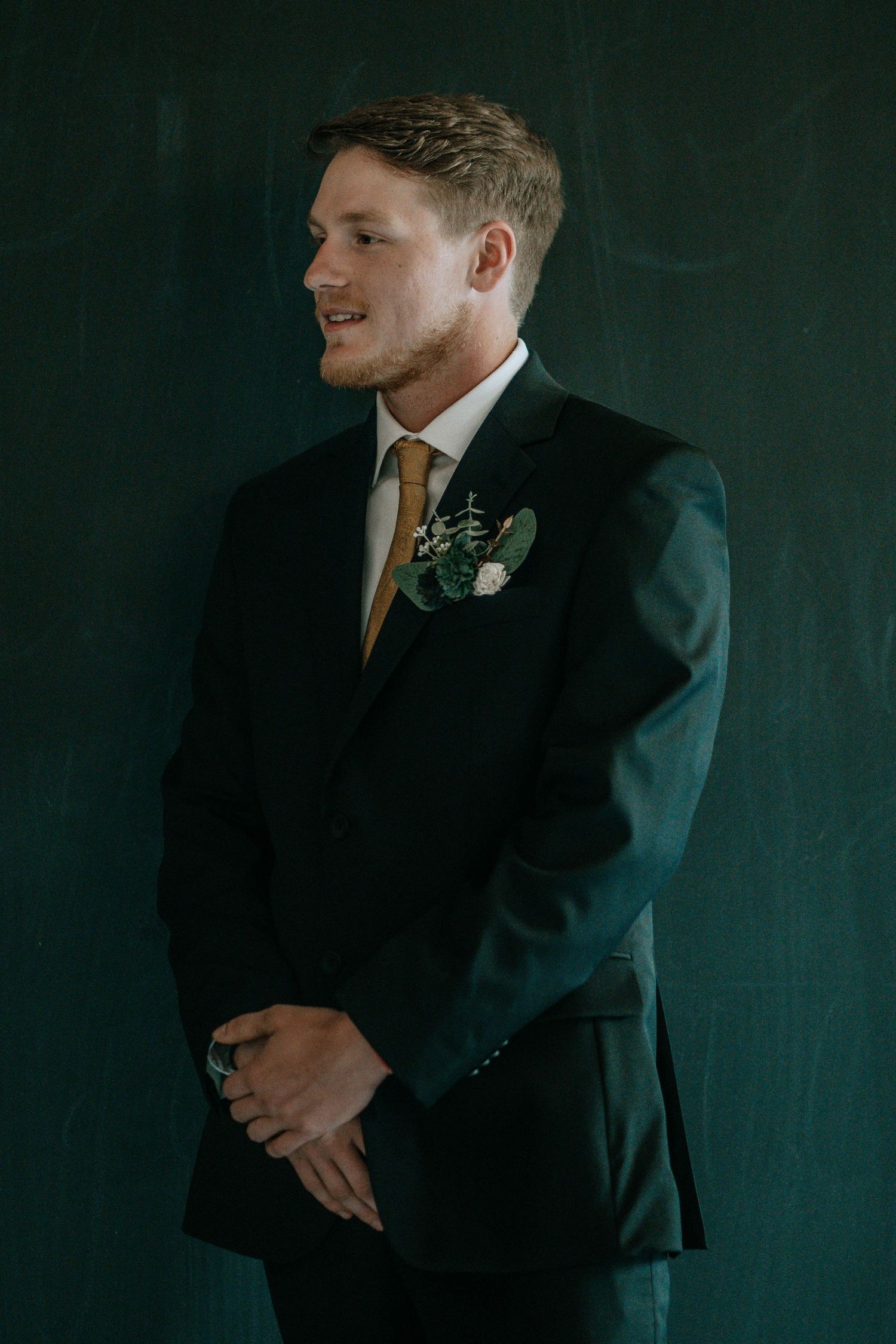 Groom in dark suit with gold tie and floral boutonniere poses confidently against a deep green background.