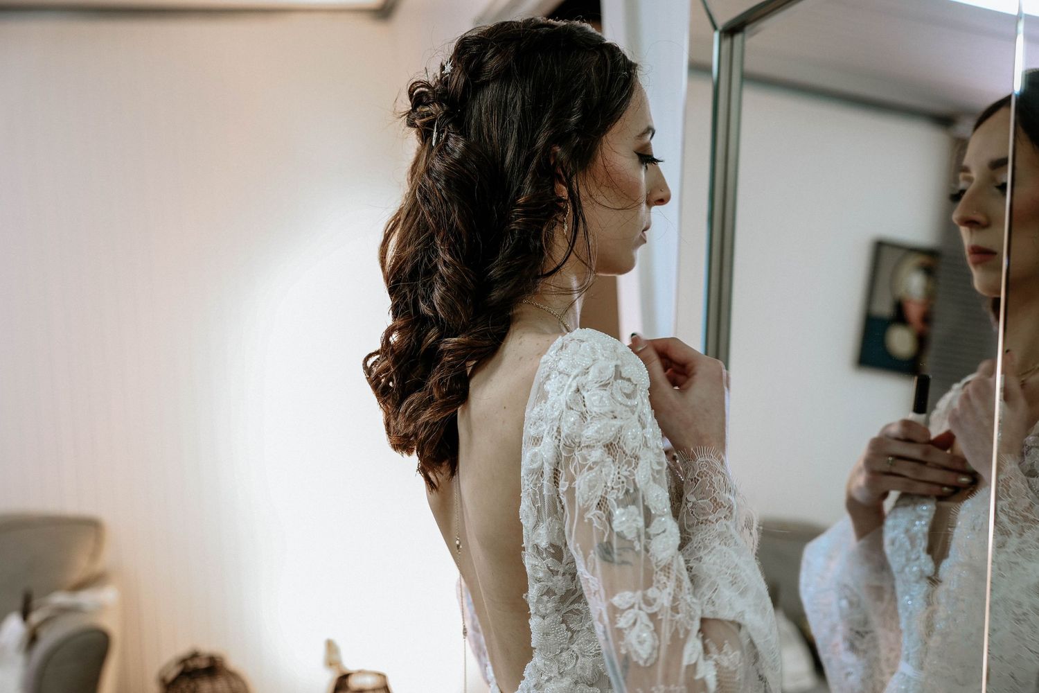 Bride in lace wedding dress getting ready, viewed from behind with elegant curled hair near a mirror.