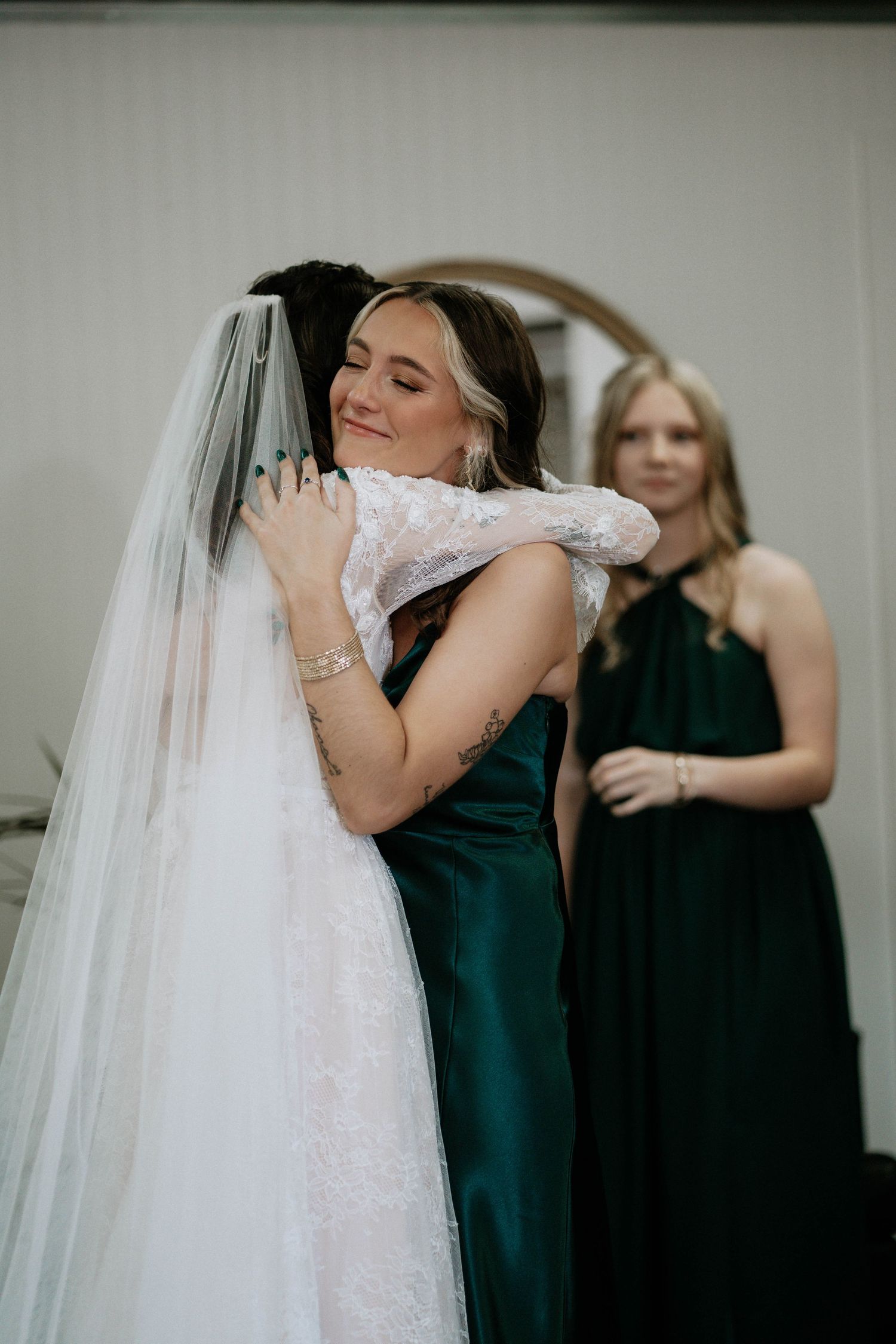 Bride in white lace gown and veil embracing bridesmaid in emerald green dress, with another bridesmaid smiling in background.